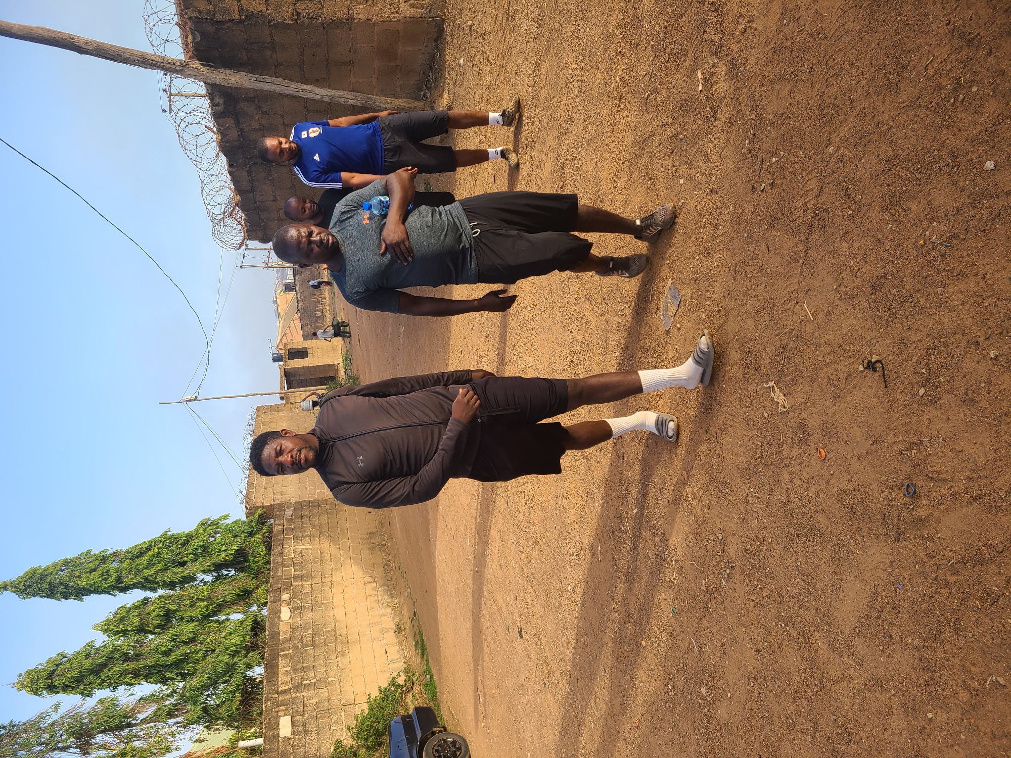 The image depicts three men standing in a dirt lot in Jos, Nigeria. The man closest to the camera wears a black jacket and shorts with white socks. The second man is wearing a grey t-shirt, black shorts, and dark colored shoes, and is holding a water bottle against his chest. The third man wears a blue soccer jersey, black shorts, and dark colored shoes. A brick wall topped with barbed wire runs along the back of the scene. A utility pole is visible near the wall. Tall trees are visible in the lower left corner. A vehicle is partially visible at the bottom of the frame. The dirt lot appears to be adjacent to residential buildings. The sky is clear and blue.