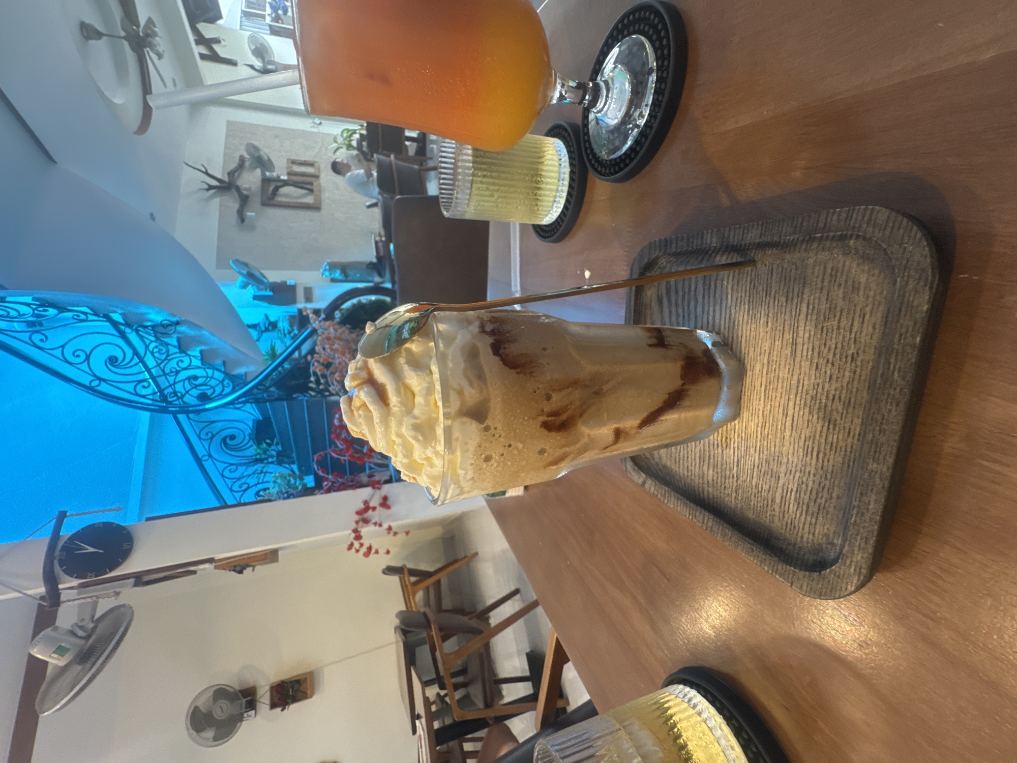 The photograph captures a close-up view of a table at an indoor cafe. Dominating the foreground is a tall glass filled with a blended beverage topped with a generous swirl of whipped cream and drizzled with caramel. A metallic spoon rests inside the glass, partially submerged. To the left, another glass, this one containing a yellow liquid, is partially visible. To the right, a tall glass with an orange-hued drink stands on a black coaster.
The table itself is made of wood, with a warm, polished surface. The background reveals the interior of the cafe, characterized by a blue-lit ceiling and a sweeping staircase with ornate metal railings. Several wall-mounted fans are visible, along with decorative elements like framed pictures and red floral accents. In the distance, a person is seated at a table, appearing to be a customer. The overall ambiance suggests a relaxed setting within a cafe located in Bac Nha Trang Ward, Vietnam.