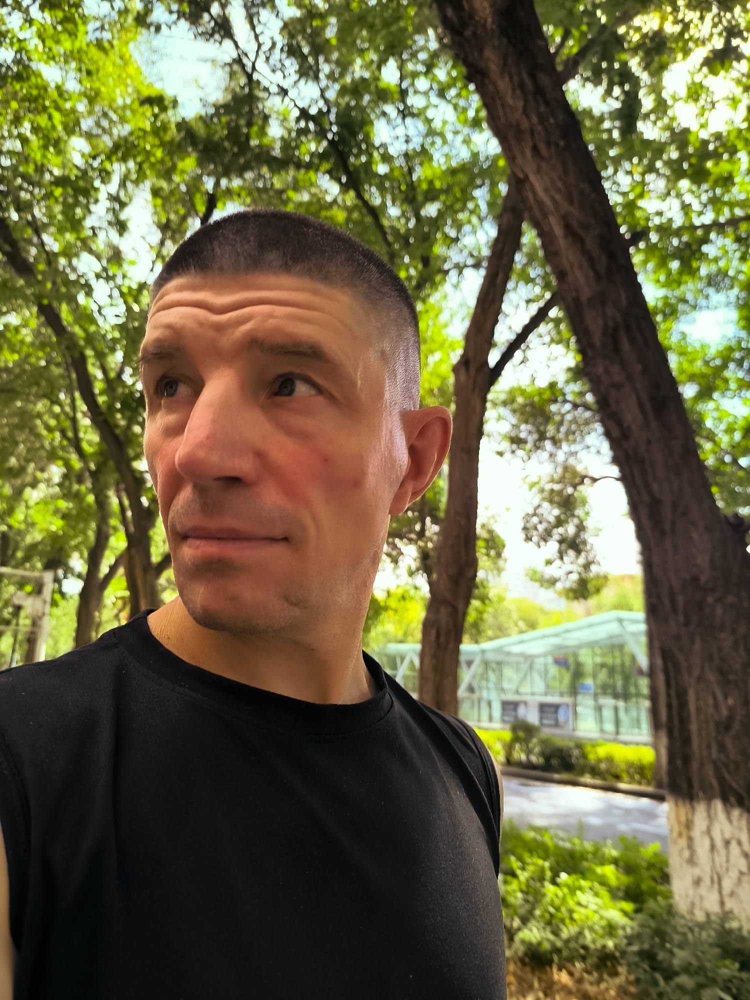 A man with a buzz cut is looking up and to his left. He is wearing a black t-shirt. He is in a park-like setting with trees and green foliage. In the background, a modern glass structure is visible, possibly a greenhouse or a transit station. The man's skin is lightly tanned and he has a light stubble on his chin. The lighting suggests it is daytime. The location is Ürümqi, China.