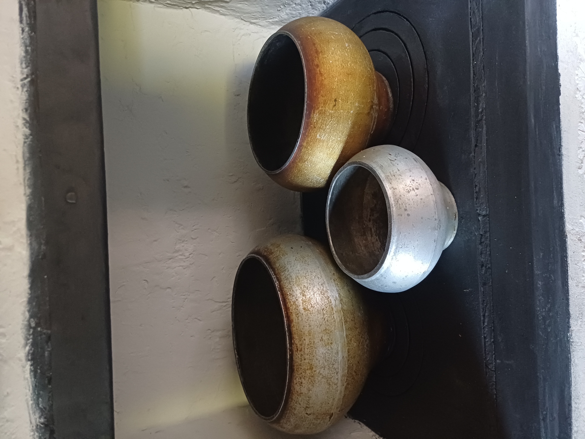 A medium shot captures three cast iron pots on a black stovetop. The pots are of varying sizes. The largest is on the left, and a slightly smaller one is on the right. A much smaller pot is positioned in front of the larger two. The pots have a rounded base and a wide opening at the top. The surfaces of the pots show signs of wear and use, with some discoloration and patina. The background consists of a textured white wall. The scene appears to be indoors, likely in a kitchen or dining area. This scene is located in Sulymivka, Ukraine.