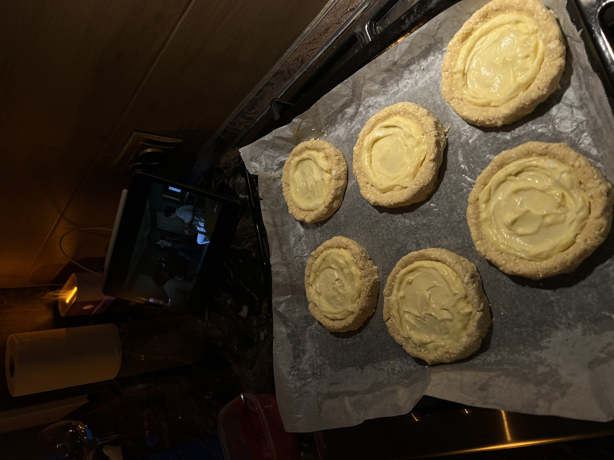 The image depicts a close-up view of six individual tarts filled with a pale yellow cream, arranged on a baking sheet lined with parchment paper. The tarts have a rustic appearance with visible crust edges. In the background, a tablet screen displays a group of people, and to the left, a glass of red liquid and a roll of paper towels are visible on a dark countertop. The setting appears to be a kitchen, likely in the city of Ternopil, Ukraine, given the provided context, though this specific location cannot be definitively confirmed from the image alone. The lighting is dim, suggesting it might be evening or night.