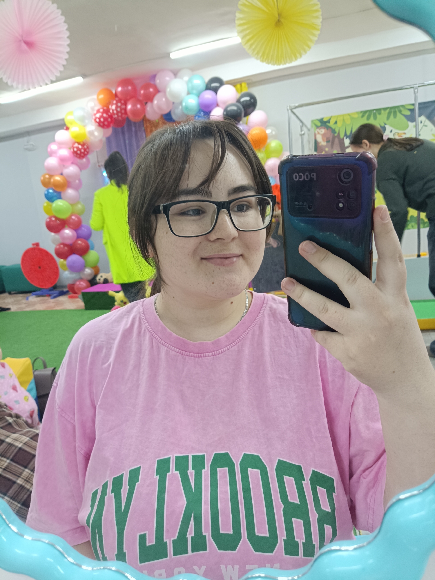 The image depicts a selfie taken by a person wearing glasses and a pink t-shirt with "BROOKLYN" printed on it. The background reveals a room decorated for a celebration, featuring a balloon arch with various colors and paper decorations hanging from the ceiling. In the background, other individuals can be seen, including one wearing a neon yellow shirt. The environment appears to be an indoor party or play area. The location is in the city of Unknown, Unknown. The selfie is taken using a cell phone in a dark blue case, which is held up in the foreground.