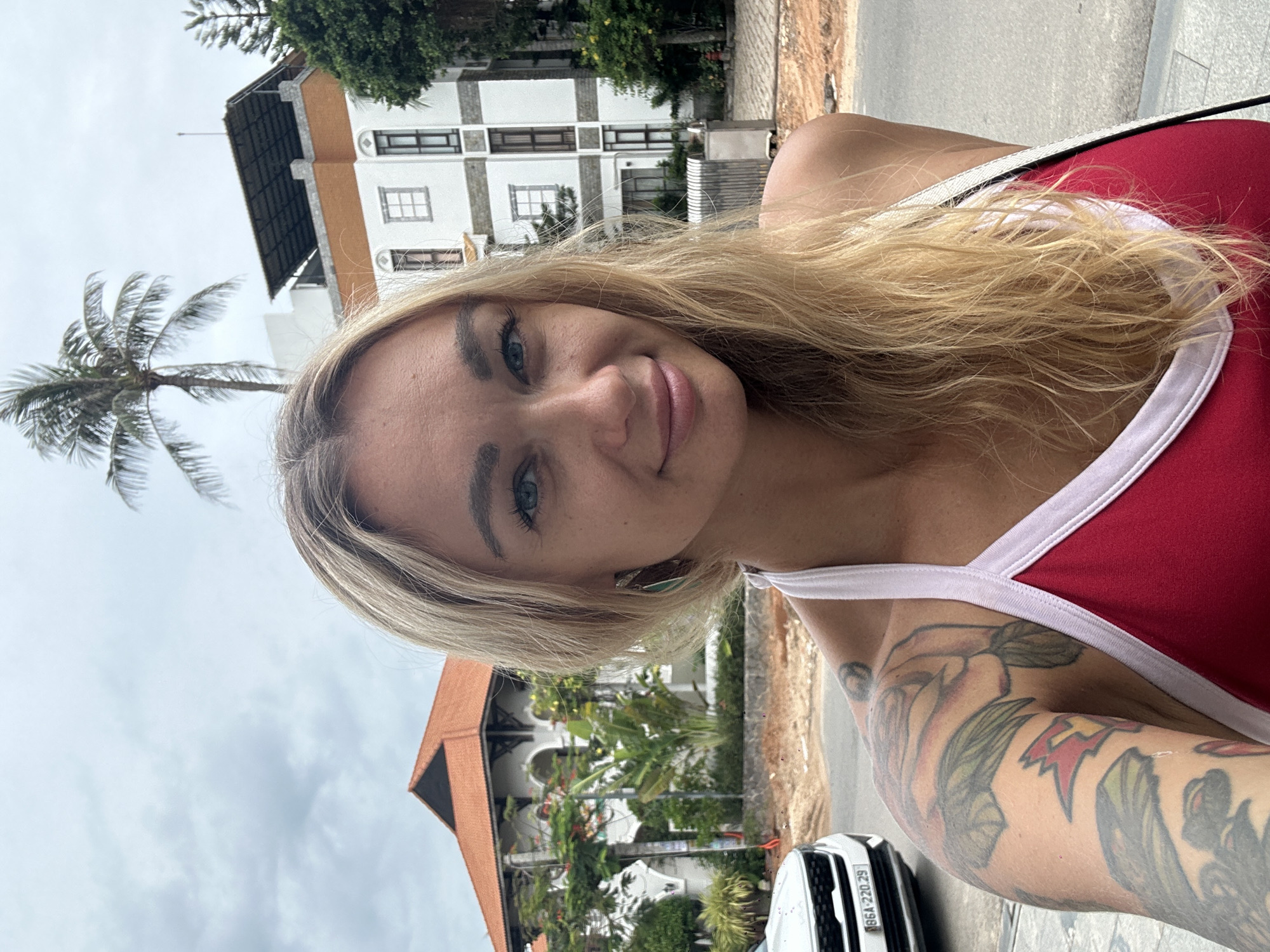 A young fair-skinned woman with blonde hair and multiple tattoos on her arm takes a selfie in Phu Thuy Ward, Vietnam. She is wearing a red tank top with white trim. In the background, there are buildings, trees, a palm tree, and a car with license plate "86A-220.29". The sky is overcast.