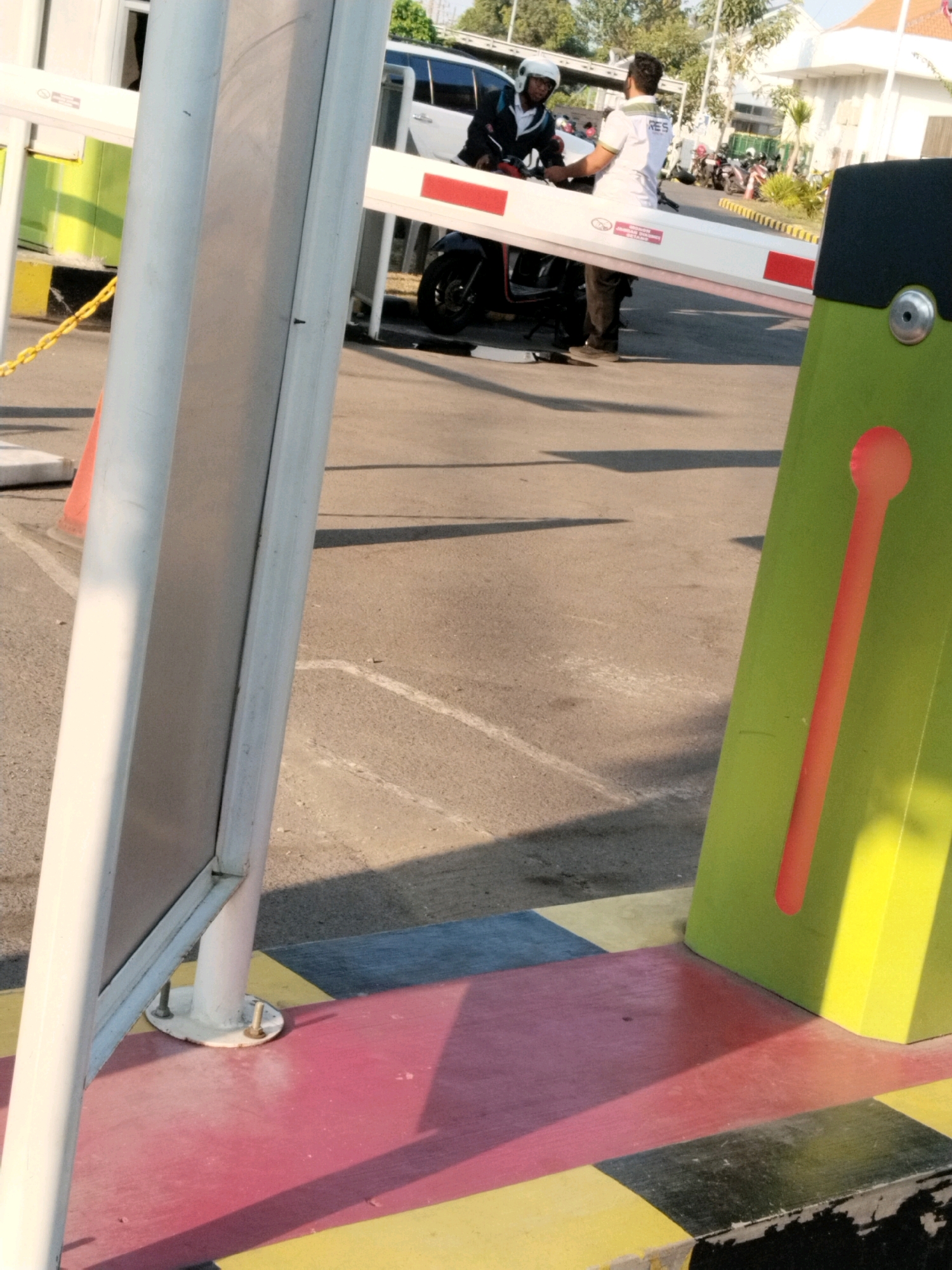 The image depicts a barrier gate at what appears to be a parking area or entrance in Wonocolo, Indonesia. In the foreground, a thick, white, vertical pole stands to the left, partially obscuring a section of a white barrier arm. The barrier arm is extended across the frame, with a red stripe visible on its surface. To the right, a bright green, angled object, likely part of the barrier mechanism, features a vertical orange stripe with a circular end. The ground is paved with asphalt, marked with white lines, and a colorful pattern of red, yellow, and black squares is visible at the base of the barrier.
In the middle ground, a person wearing a white helmet and a dark jacket is seated on a motorcycle. They are interacting with another person, who is wearing a white shirt with green trim and black pants. The person in the white shirt is standing next to the motorcycle. Behind them, several other motorcycles are parked, and a white SUV is visible. In the background, buildings and trees suggest an urban or semi-urban environment. The lighting indicates it is daytime, with shadows cast on the ground.