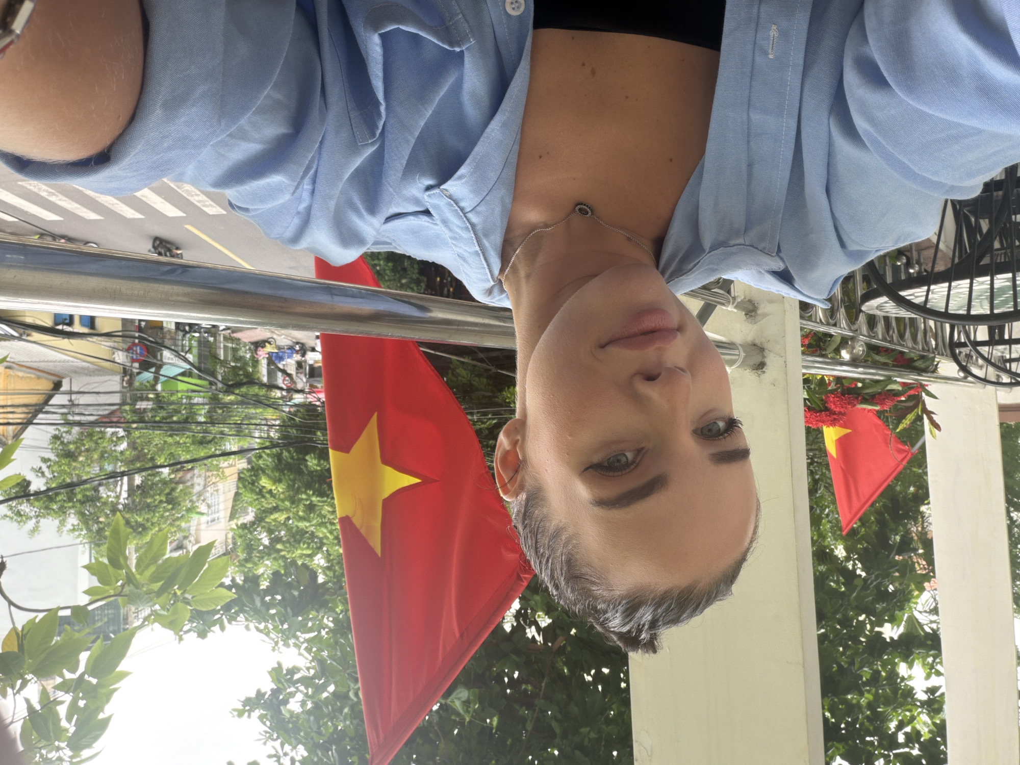 The original image is oriented sideways, appearing as if the subject is upside down, taking a selfie. In the foreground, a person, likely female, is partially visible from the chest up. She has light skin, dark eyebrows, and blue or grey eyes, looking towards the camera with a neutral to slightly contemplative expression. Her hair is dark and styled back. She wears a light blue button-down shirt unbuttoned over a black top, and a delicate silver necklace with a small dark charm.
A metallic railing runs horizontally across the mid-ground, separating the person from the background. Beyond the railing, a street scene unfolds, visible from an elevated perspective. A zebra crossing and a yellow dividing line are distinguishable on the road, where small vehicles, likely scooters or motorbikes, are present.
The background is dominated by lush green trees and foliage. Prominently displayed are at least two Vietnamese flags, red with a yellow star, hanging from structures. Various urban buildings are nestled among the trees. Overhead, utility wires crisscross the sky. The bright daylight suggests it's daytime. The setting is an outdoor urban environment, consistent with Nha Trang Ward, Vietnam. No discernible text is visible.