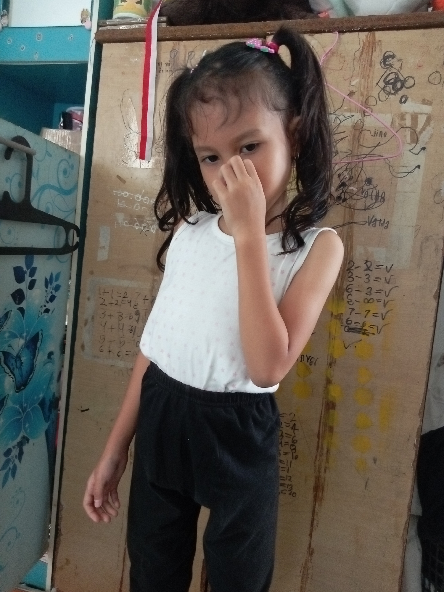 A young girl with dark hair pulled into two pigtails, wearing a white sleeveless shirt with pink polka dots and black sweatpants, stands in front of a wooden panel covered in handwritten math problems and drawings. She is touching her face with her right hand, her expression is neutral. The background appears to be an indoor setting, possibly a bedroom or play area, with a blue cabinet on the left featuring a butterfly motif. The wooden panel has numerous equations like "1+1=2", "2+2=4", "3+3=6", "4+4=8", "5+5=10", "6+6=12", "7+7=14", "8+8=16", "9+9=18", and "10+10=20", as well as some scribbled drawings and what appears to be a pink clothes hanger. The environment suggests a casual, possibly educational, home setting. Given the general context and typical domestic scenes in Indonesia, it is plausible this scene is located within a residential area in Batam, Indonesia.