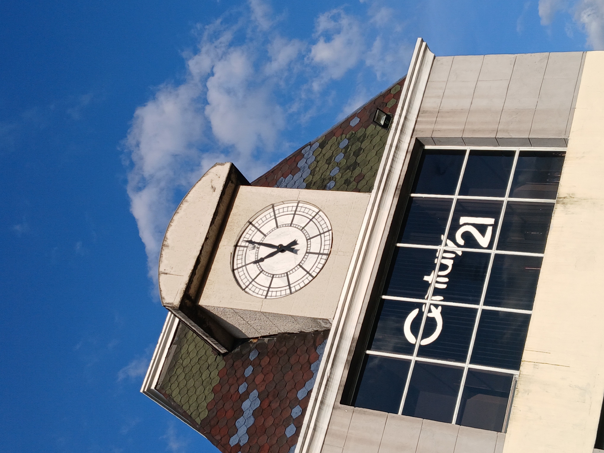 The image captures a low-angle view of a building in Guayana City, Venezuela, under a partly cloudy blue sky. The building features a clock tower with a visible clock face showing a time around 8:49. The roof of the tower is covered in multi-colored tiles. Adjacent to the clock tower is a multi-pane window with the "Century 21" logo displayed. The building's exterior is a mix of grey and light beige concrete. There are no people visible in the frame. The angle suggests the photo was taken from street level.
