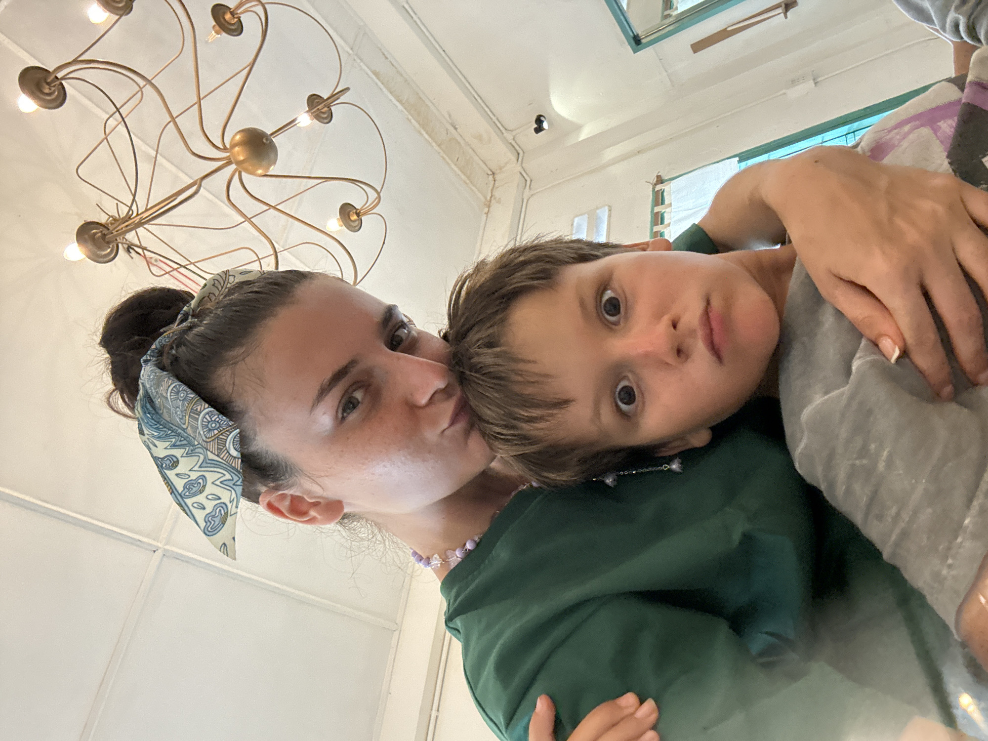 A low-angle, close-up, selfie-style photograph captures a woman and a young boy in an indoor setting. The woman, positioned behind and slightly above the boy, has her hair pulled up in a bun and is wearing a blue and white patterned bandana. She is leaning in, with her forehead touching the boy's head, and her left arm is wrapped around his shoulders. She is wearing a green long-sleeved shirt and a beaded necklace. The boy, in the foreground, has wide, surprised eyes and a slightly pursed mouth. He is wearing a grey t-shirt. The background features a white, paneled ceiling with a decorative brass chandelier. The overall lighting suggests daylight, possibly from windows that are not directly visible. There are no visible contextual clues to indicate the location within Phu Thuy Ward, Vietnam, or any specific city.