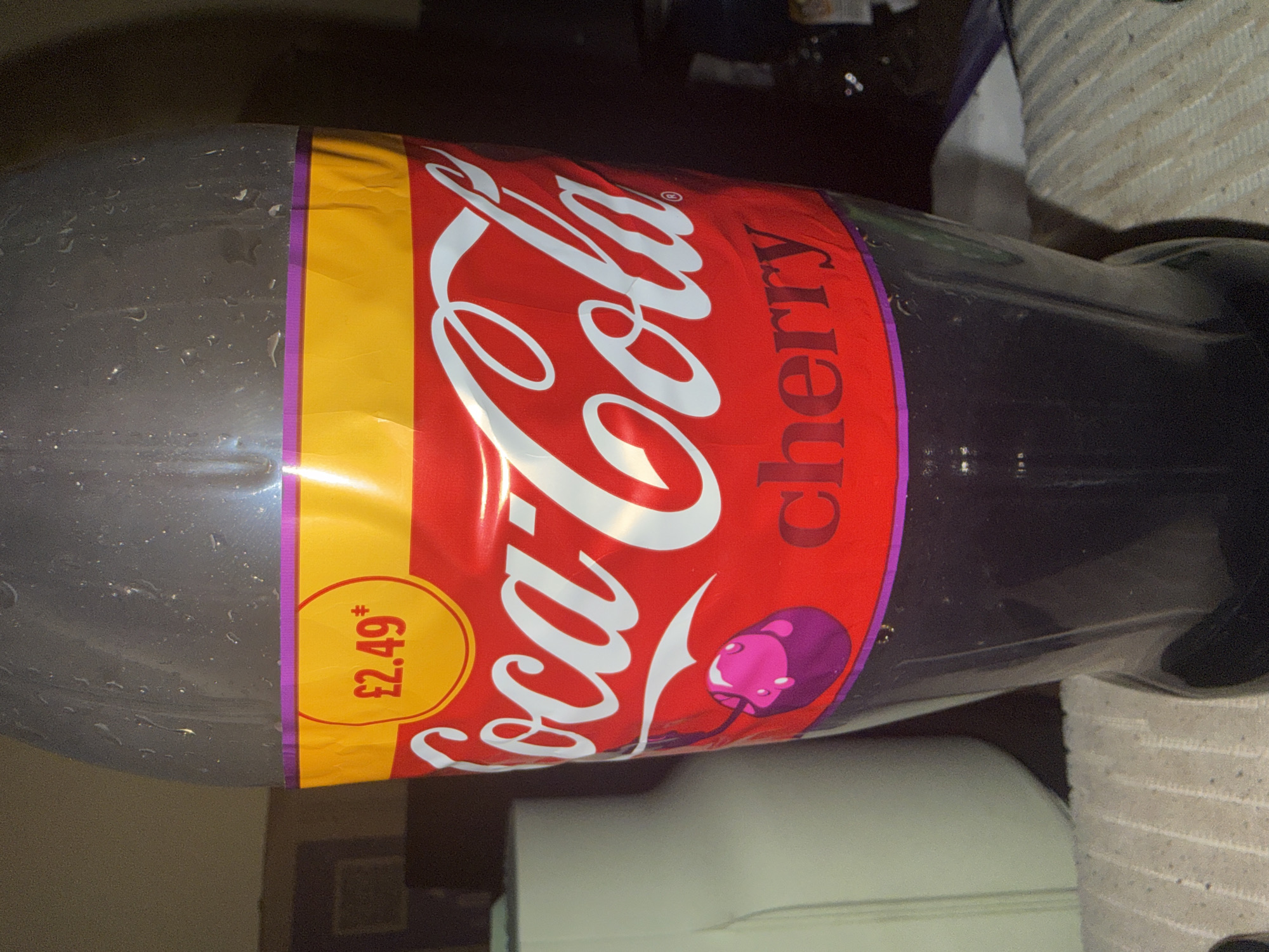 A close-up shot features a plastic bottle of Coca-Cola Cherry soda. The bottle's label is predominantly red with the iconic white Coca-Cola logo. Above the logo, a yellow band displays the price "£2.49" in a red circle. Below the logo, the word "cherry" is printed in a darker red. A graphic of a pink and purple cherry with a stem is also visible on the label. The bottle is filled with a dark purple liquid and appears to have condensation droplets on its surface. The background is dark and out of focus, with hints of furniture and a light-colored textured surface, possibly a table, in the foreground. The image does not contain any specific landmarks or indications of a location in Cheltenham, United Kingdom.
