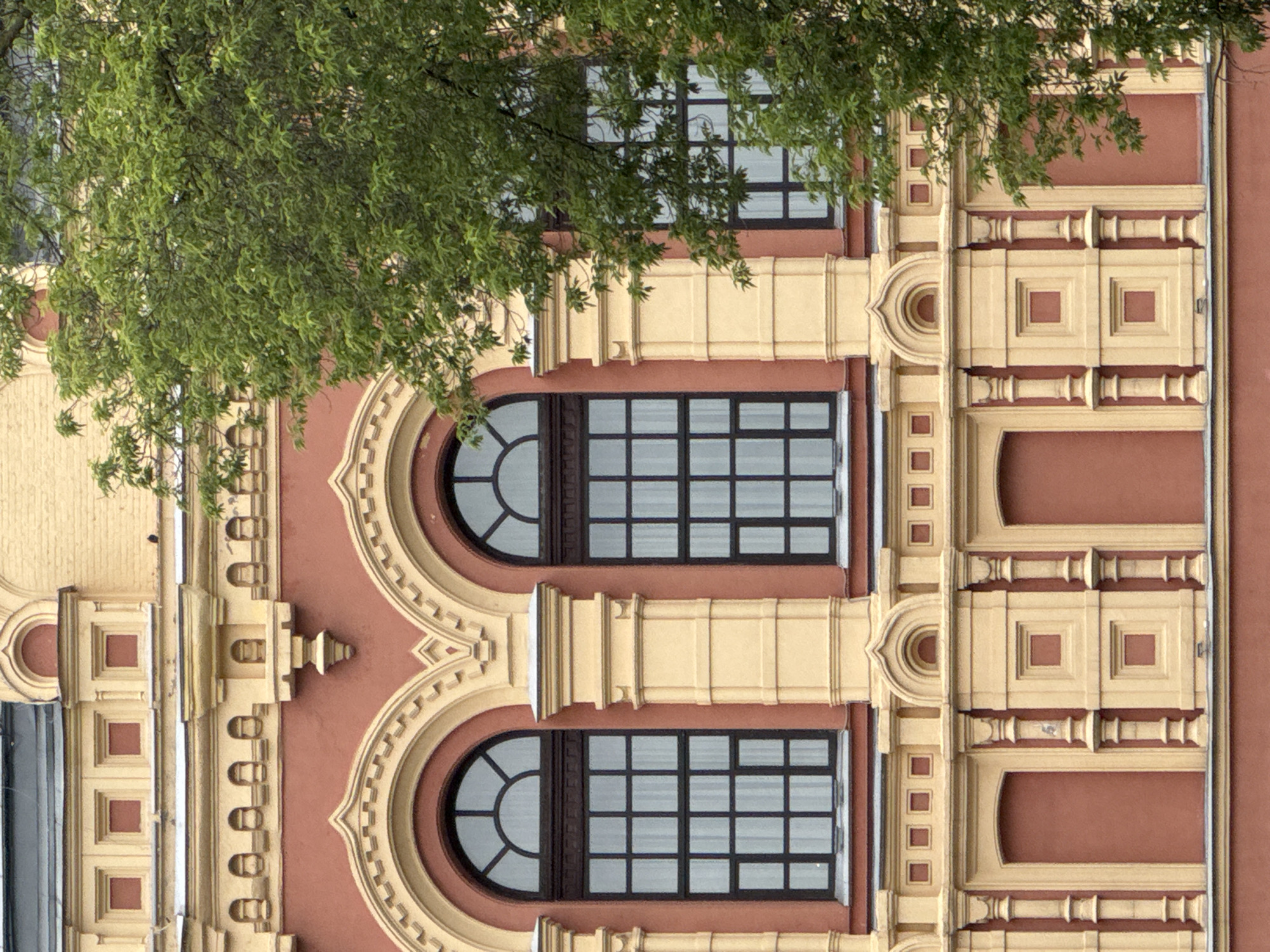 The image shows a close-up of the facade of a building, likely in Nizhny Novgorod, Russia, given the architectural style. The building has a reddish-pink stucco exterior with ornate yellow decorative elements. There are at least two large, arched windows visible, each with multiple panes and dark frames. Above the windows, there are more decorative architectural features, including what appear to be blind arched recesses and square indentations. Below the main windows, there are several smaller arched openings with solid reddish-pink infills, possibly indicating former doorways or windows that have been covered. A large tree with lush green foliage partially obscures the right side of the building. The architectural details suggest a historical building, possibly from the late 19th or early 20th century.