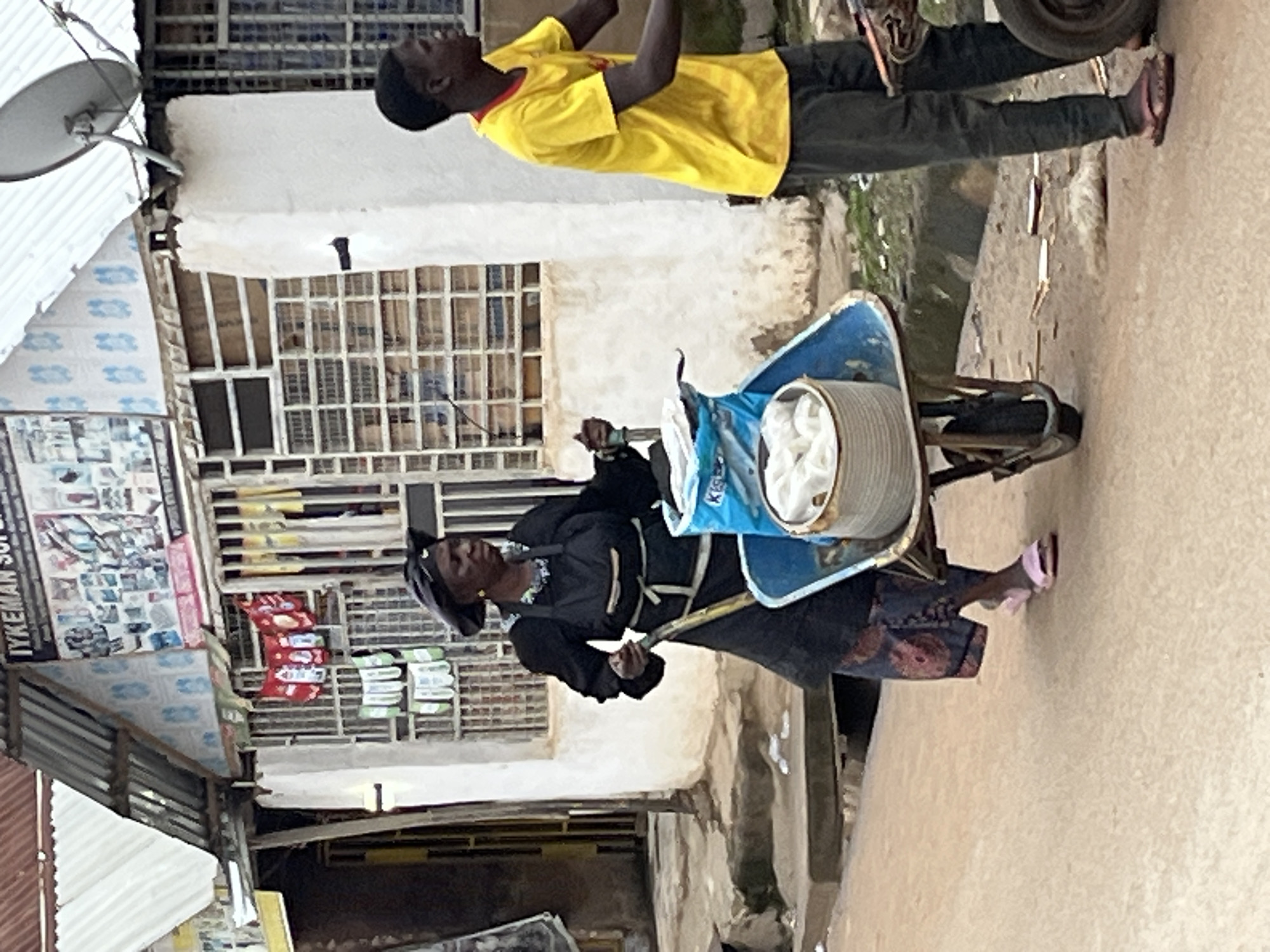 In Abuja, Nigeria, on a dusty street, a woman dressed in a black apron and headscarf pushes a wheelbarrow filled with goods. She wears a patterned skirt and pink sandals. To her right, a young man in a yellow t-shirt and dark pants walks alongside. In the background, a building displays signs and has barred windows. The scene suggests a busy marketplace or street vendor area.