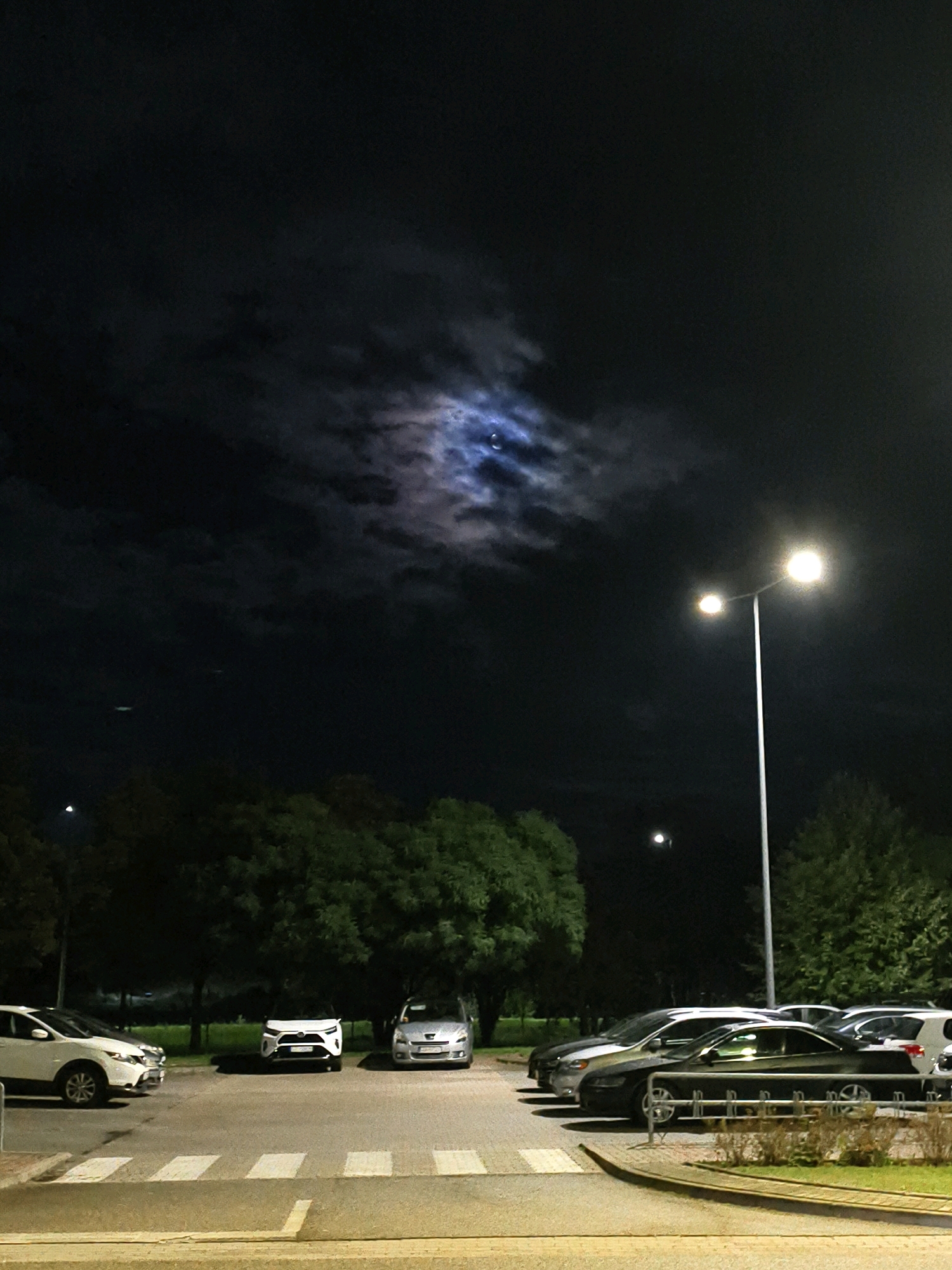 The image depicts a nighttime scene in a parking lot, possibly in the city of Tczew, Poland. The sky is dark with clouds partially obscuring a bright moon. A street lamp with two lights illuminates the area. Several cars are parked in rows on an asphalt surface marked with white lines. In the foreground, a pedestrian crossing is visible. Trees line the background.