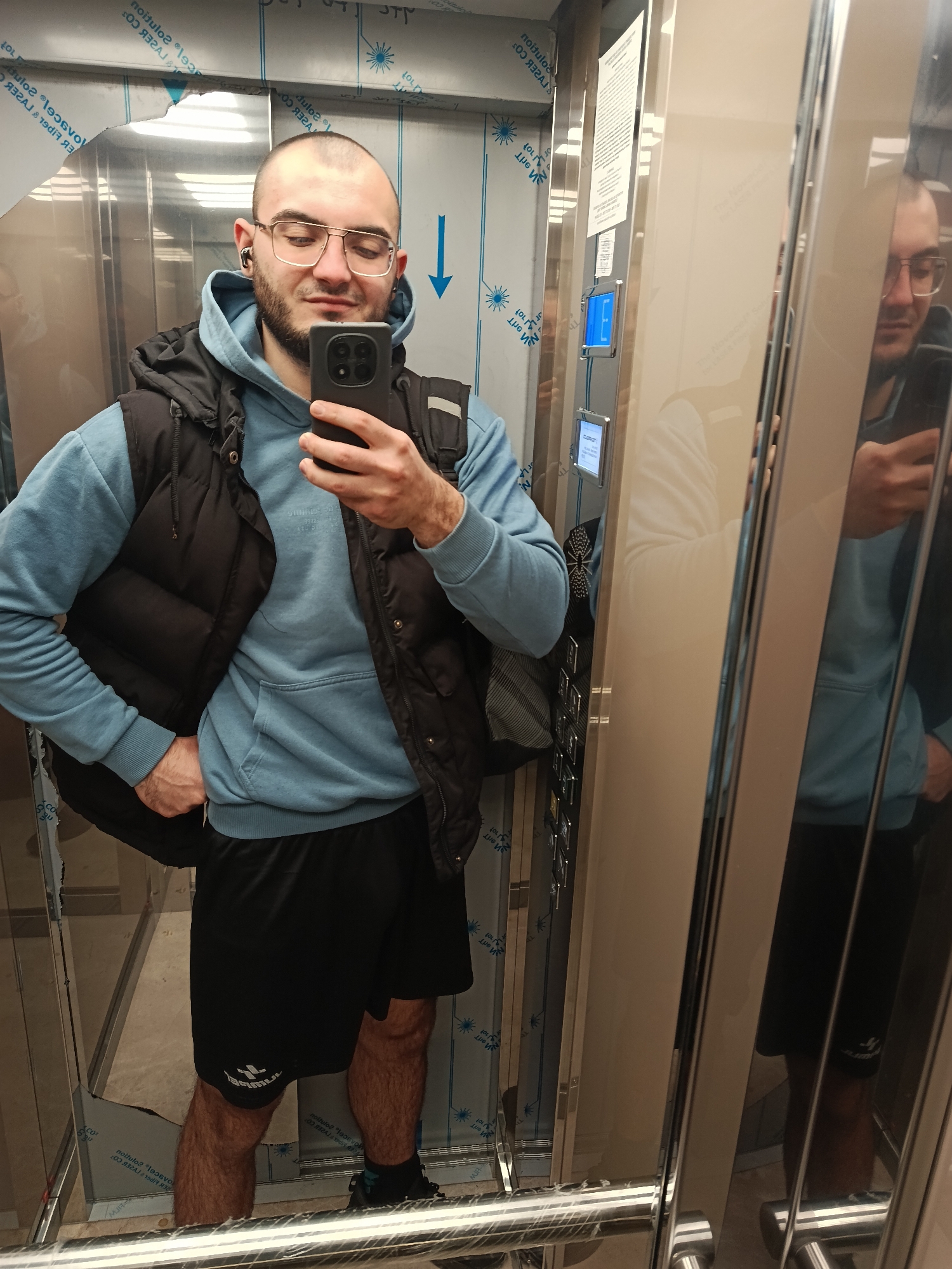 A man is taking a selfie in an elevator. He is fair-skinned, bald on top with short dark hair on the sides, and has a dark beard. He wears wire-rimmed glasses, an earbud in his left ear, a light blue hooded sweatshirt, a black puffy vest, and black shorts. He holds a black cell phone in his right hand, taking the picture. He has his left hand on his hip. The background of the elevator shows stainless steel walls and a control panel on the right side. His reflection is visible in the mirror on the right. There are no clear indicators of the location in Plovdiv, Bulgaria.