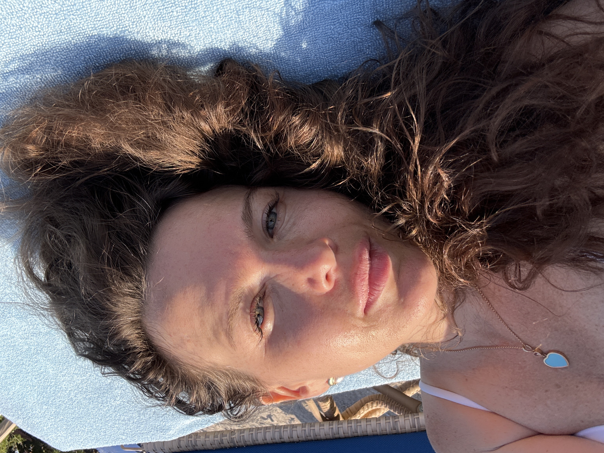 A woman with long, dark brown, curly hair is lying on a blue towel, possibly outdoors. Her face is angled towards the camera, with her eyes looking slightly to the left. She has fair skin, light blue eyes, and her lips are slightly parted. A strand of her hair falls across her forehead and cheek. She is wearing a white bikini top and a delicate gold necklace with a heart-shaped pendant containing a light blue stone. The lighting suggests it is daytime, with sunlight casting shadows. The background shows a woven chair and a bright blue towel. There is no indication of the city of Dubai or the United Arab Emirates in this image.