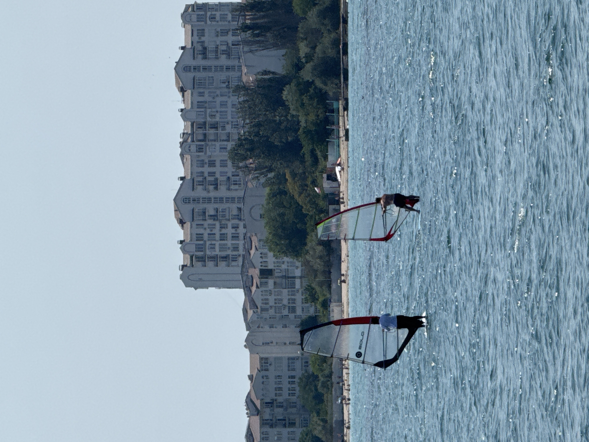 In Sevastopol, Ukraine, two individuals are windsurfing on a bright, sunny day. The sea is a vibrant blue with visible ripples and occasional glints of sunlight. In the foreground, a windsurfer with a white shirt and dark pants is visible behind a sail with black and red trim. To the right, another windsurfer, wearing dark shorts, is partially obscured by a sail with red trim.
The middle ground of the image features a beach populated with numerous sunbathers and umbrellas. Beyond the beach, a line of lush green trees forms a backdrop. Dominating the background is a large, multi-story white building with numerous windows and balconies, characteristic of an apartment complex or hotel, set against a pale, cloudless sky.