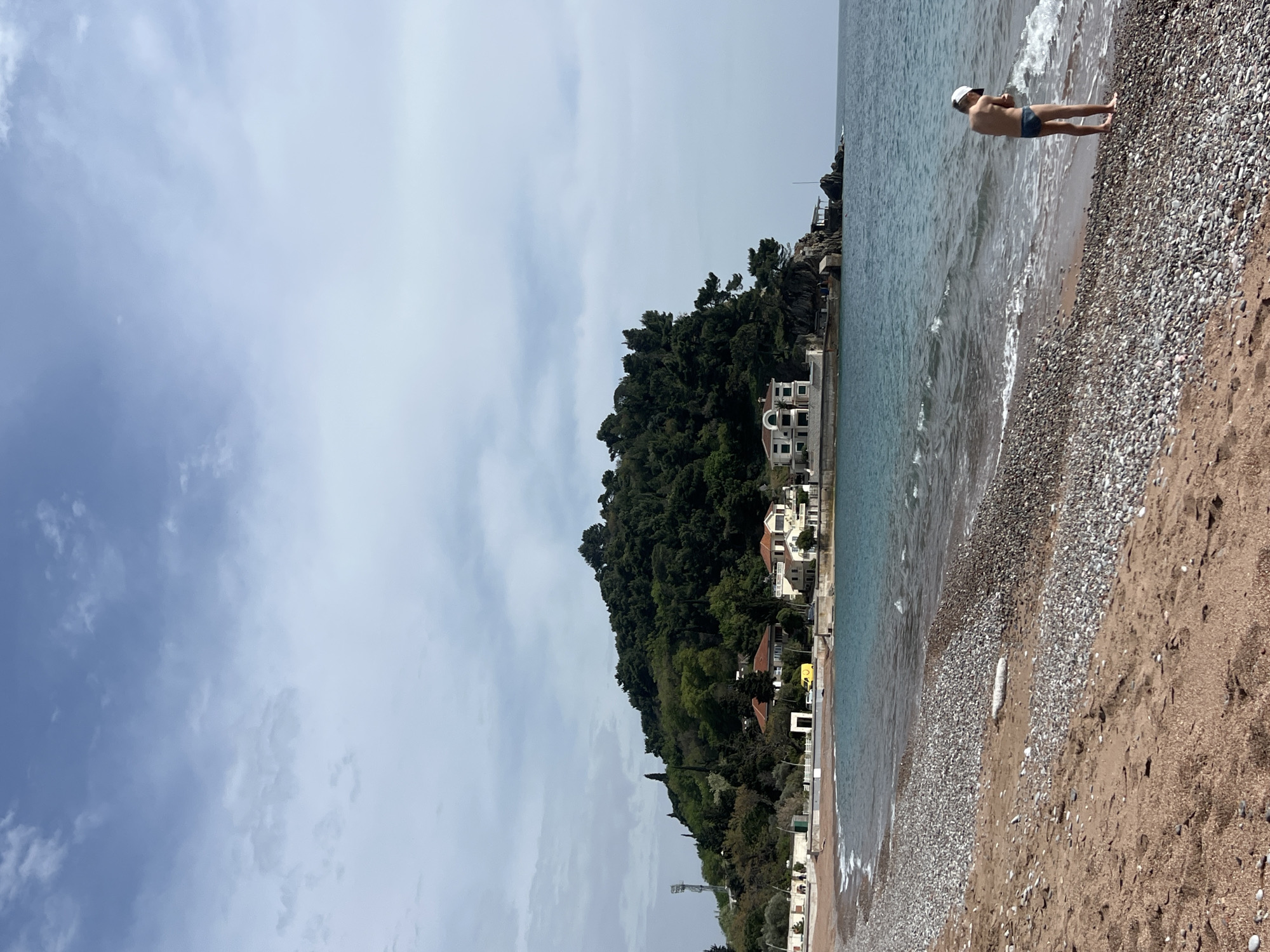 The image depicts a serene coastal scene, likely in Petrovac, Montenegro, given the provided context. The perspective is from the beach, looking towards the sea and a forested hillside.
In the foreground, a sandy and pebbly beach stretches across the lower portion of the frame. Gentle waves lap at the shore, where a single person stands. This individual is seen from behind, wearing a white cap and dark blue swim briefs, with the water reaching approximately to their knees. The beach transitions from larger pebbles near the waterline to finer sand further back.
The midground features the calm, blue-green waters of the sea with small, soft waves. Beyond the water, a prominent, verdant hill rises, densely covered with green trees and foliage. Nestled along the base of this hill and extending to the waterfront are several multi-story buildings, predominantly white or light-colored with red or orange-tiled roofs, characteristic of Mediterranean architecture. Some concrete structures or a promenade are visible at the water's edge in front of these buildings. A yellow vehicle is partially visible among the buildings on the left side.
The sky, forming the background, is partly cloudy, with large expanses of light gray and white clouds, through which patches of lighter blue sky are visible. The overall lighting suggests it is daytime, possibly late morning or early afternoon. The scene appears calm and quiet, with no obvious signs of intense activity or strong emotions. No discernible text is visible in the image.