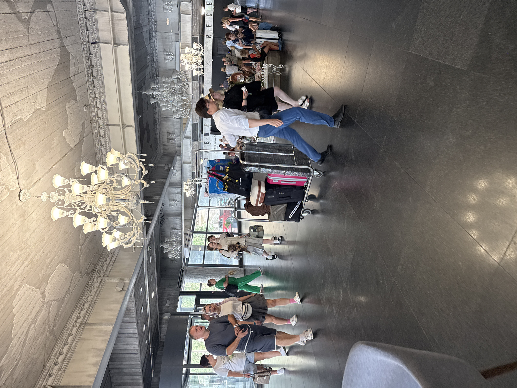The image depicts the interior of a hotel lobby. The architecture features a high, ornate ceiling with decorative moldings and large, elaborate chandeliers. The floor is a dark, polished tile. Several individuals are present, most of whom are walking or standing with luggage. In the foreground, a man in a white shirt and blue pants is pushing a luggage cart laden with several suitcases. To his left, a woman in a black dress and hat is walking beside him. Further back, a group of people are scattered throughout the lobby, some conversing, others moving with their belongings. A woman on the left is walking with a small cat in a carrier. The overall impression is of a busy, possibly transitional space within a hotel. The location is identified as Yalta, Ukraine.