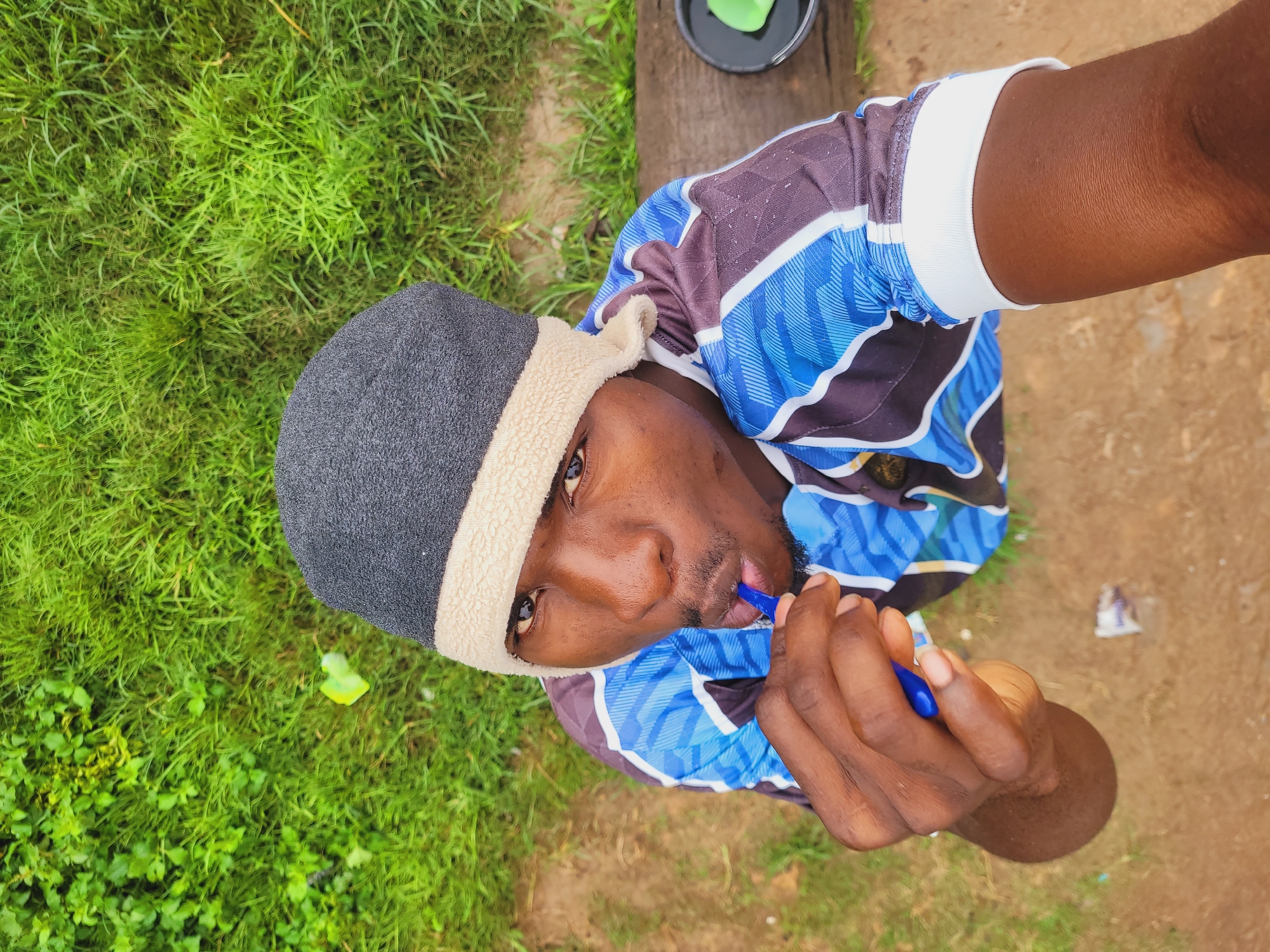 A high-angle, medium shot captures a young Black male from the chest up, wearing a blue and black patterned t-shirt and a gray beanie with a cream-colored trim. He is holding a blue object, possibly a toothbrush, to his mouth as if brushing his teeth. The subject is outdoors, with a background of lush green grass and a wooden surface to the right. The ground appears to be dirt, with some scattered litter. The lighting suggests it is daytime, with soft shadows. The overall scene is casual and appears to be in a residential or natural setting within the city of Bukuru, Nigeria.
