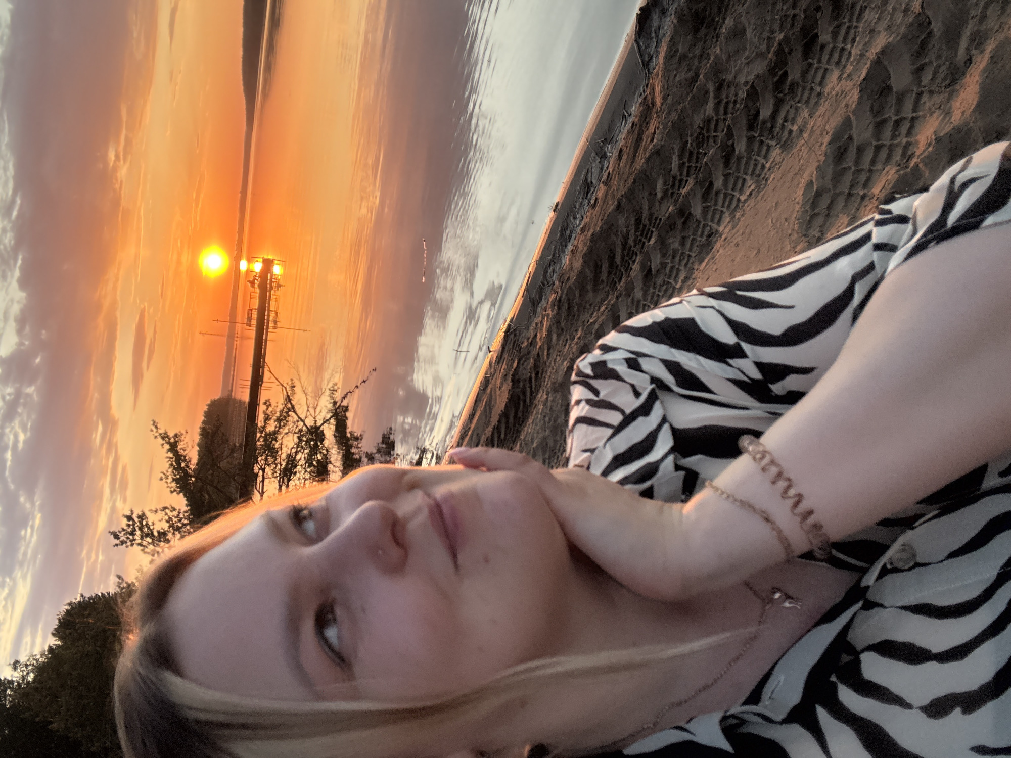 A young woman is photographed at sunset on the bank of the Kama River in Chaykovsky, Russia. The woman, who has blond hair, holds her face in her hand. She wears a black and white zebra-striped shirt and several bracelets and a necklace. In the background, a utility pole rises out of the water with an orange sky. The river reflects the sunset. The bank is rocky.