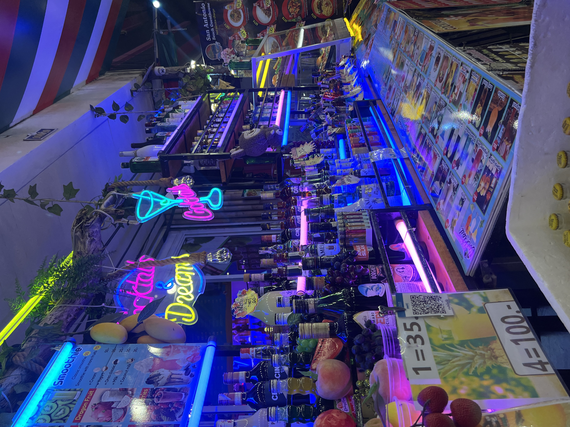 This is a photograph of a vibrant cocktail and smoothie bar located in Karon, Thailand. The bar is brightly lit with neon signs, including a pink sign that reads "Cocktails & Dreams." The shelves behind the bar are stocked with numerous bottles of alcohol and other beverages. In the foreground, there is a display of fresh fruit, including mangoes and lychees. A menu board is visible with prices for drinks, with one item listed as "1 = 35" and another as "4 = 100.-". The overall atmosphere is lively and inviting, typical of a tourist hotspot in Karon. There are no people visible in the frame.