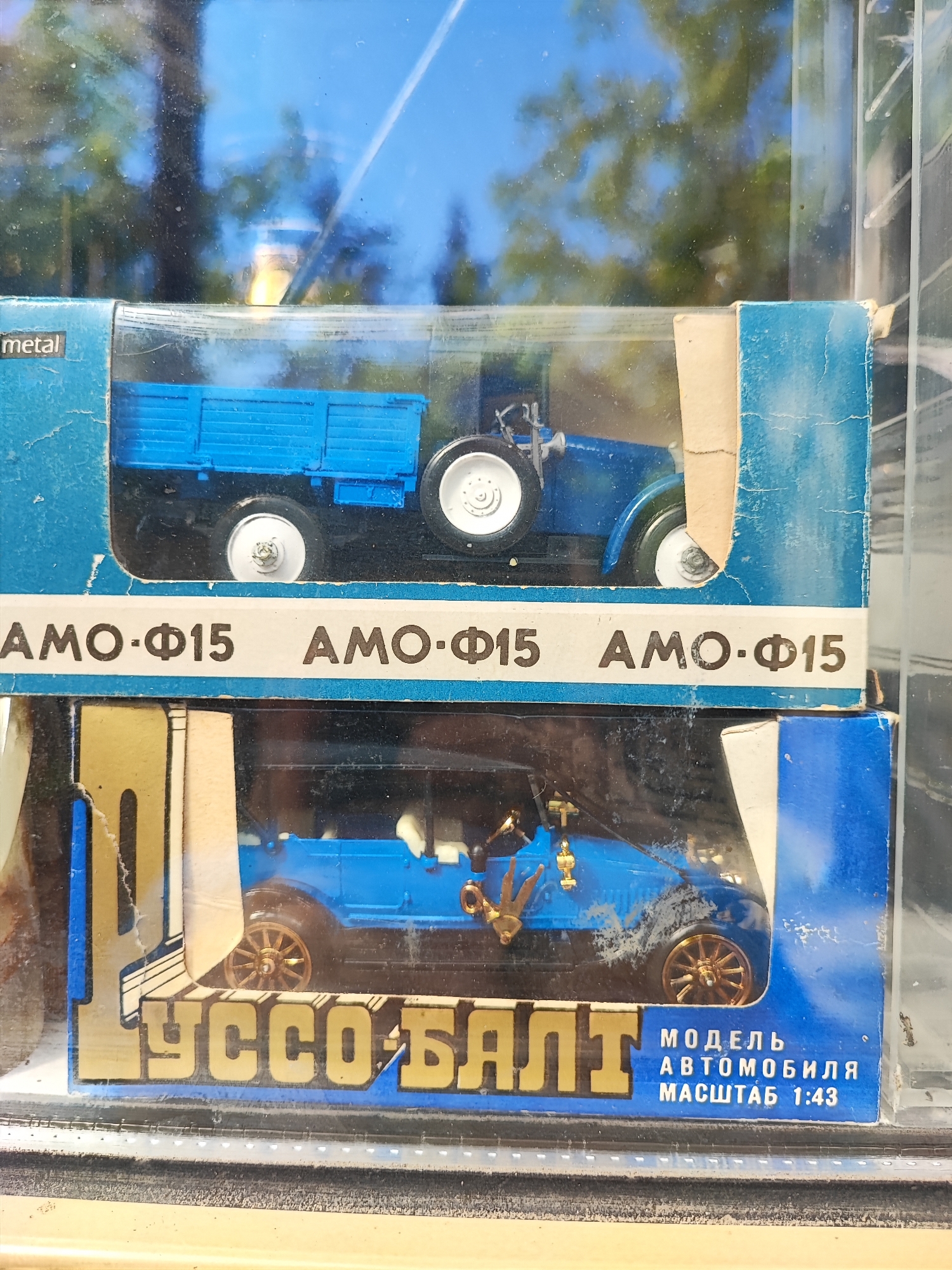 The image shows two blue toy trucks displayed in clear plastic packaging. The top truck is a flatbed truck and the bottom truck is an open-top vehicle. Both trucks are branded with Russian text and appear to be scale models. The packaging is blue with yellow accents. The reflections in the packaging suggest the display is likely inside a store or museum. Without further information, the specific location within Pyatigorsk, Russia, cannot be determined.