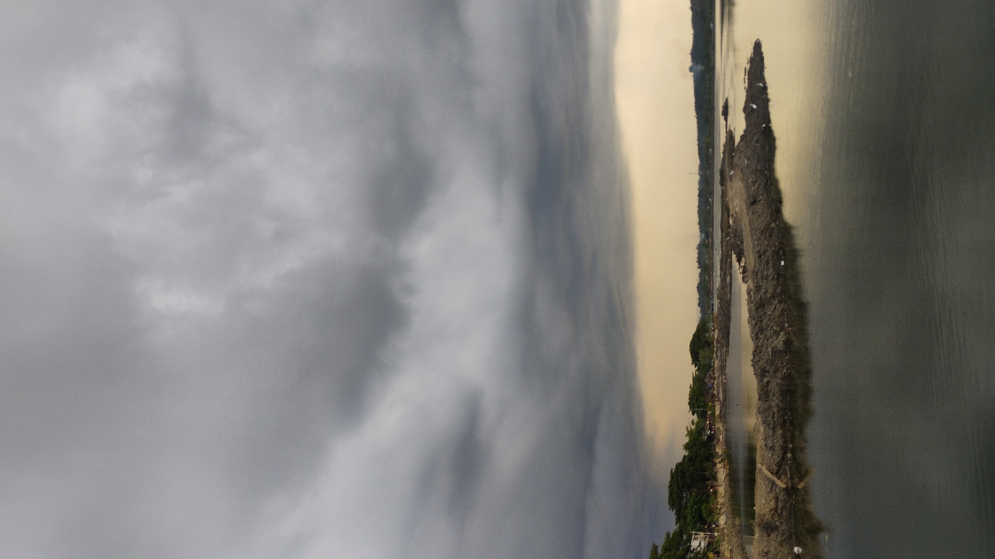 The image depicts a body of water, likely a river or bay, under a dramatic sky filled with heavy, dark gray clouds that dominate the upper portion of the frame. The clouds suggest an impending storm or overcast weather. The water's surface is relatively calm, reflecting the muted light and the dark clouds above.
In the middle ground, a narrow, man-made embankment of dark soil protrudes into the water. This embankment is irregular in shape and appears to be part of some construction or land reclamation project. Scattered on this embankment are several small white birds, possibly egrets or herons, standing still.
Along the far bank of the water, a line of trees forms a natural border, with some signs of human habitation visible behind them, including small structures and what appear to be makeshift buildings or shacks. The distant shoreline is lined with more vegetation and hints of buildings, suggesting a populated area.
The lighting is subdued, with a yellowish-tan hue on the horizon, possibly indicating the sun trying to break through or reflecting off the atmosphere. The overall scene conveys a sense of atmospheric tension and a rural or semi-urban coastal environment. Given the context, this scene could be representative of areas near the coast in Lhokseumawe, Indonesia, where such landscapes with bodies of water, embankments, and distant settlements are common.