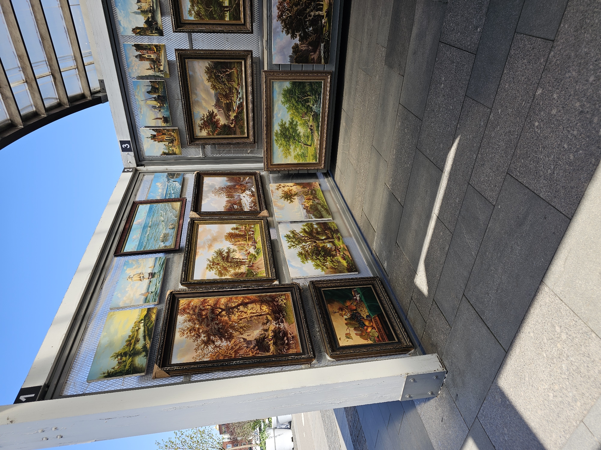 A collection of framed paintings is displayed on a metal rack outdoors. The rack appears to be part of an art market or exhibition, likely in a public space in Moscow, Russia. The paintings depict landscapes and still lifes. The environment is a city sidewalk with shadows cast from the overhead structure. No people are visible in the immediate frame.