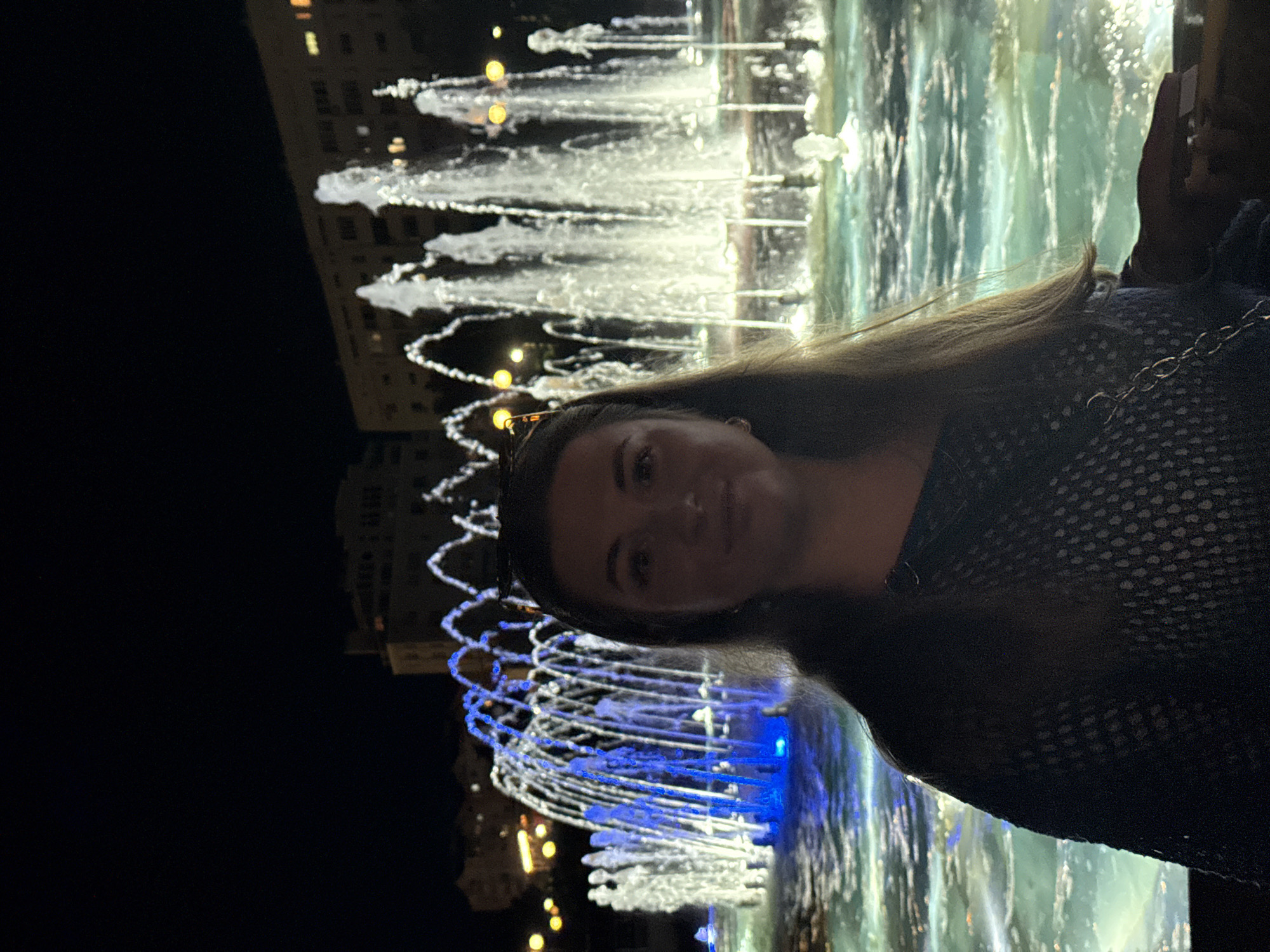 A full-frame, eye-level, medium shot shows a young woman with medium-length brown hair, wearing a dark blue, patterned top. She is positioned in the center of the frame, looking directly at the camera. Behind her, a large fountain is illuminated with blue and white lights. The water from the fountain is spraying upwards and outwards, creating a dynamic effect. In the background, buildings are visible, some with lights on. The scene is set at night, indicated by the dark sky. The woman's expression is neutral, and her body language is relaxed. The focus is on the woman, but the fountain and the background also contribute to the overall ambiance of the scene.