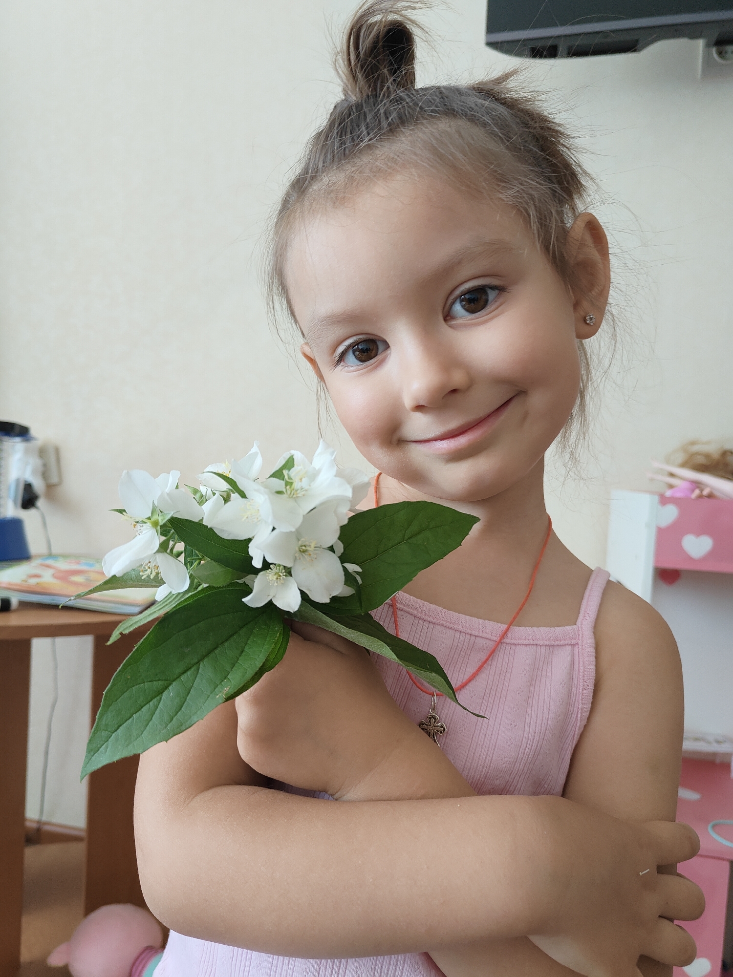 A young girl with light brown hair, pulled into a messy bun on top of her head, is captured in a close-up indoor portrait, smiling gently at the camera. She has bright, dark brown eyes and a fair complexion. She is wearing a light pink, ribbed sleeveless top and a thin orange cord necklace with a silver-colored cross pendant. A small, sparkling stud earring is visible in her right ear.
She holds a fresh bouquet of white jasmine flowers with dark green leaves close to her chest, her arms crossed. The vibrant white flowers contrast with her pink top and the green foliage.
The background suggests a casual home environment. On the left, a portion of a light wooden table is visible, holding what appears to be a book or coloring book, and possibly other household items. To the right, a segment of a pink and white shelving unit, decorated with white heart cutouts, is partially seen. A dark, rectangular object, possibly a wall-mounted television or monitor, is in the upper right background. The walls are a neutral light beige color. The lighting is soft and even, characteristic of indoor ambient light. The girl's expression is one of calm happiness and innocence. No discernible text is visible in the image. The scene is set in Chornomorsk, Ukraine.