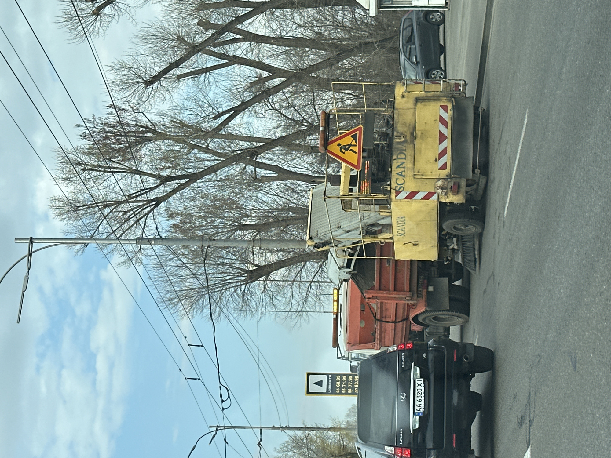 The image captures a street scene in Kyiv, Ukraine, featuring road maintenance. A yellow and orange Scania truck with a "work in progress" sign is parked along the road. Behind it is a black Lexus GX470 with the license plate "AA 6320 XI." A fuel price sign for Ukrnafta is visible, showing prices for 95, 95, 90, and diesel fuel. Bare trees line the roadside, and overhead power lines stretch across the sky. The sky is partly cloudy.