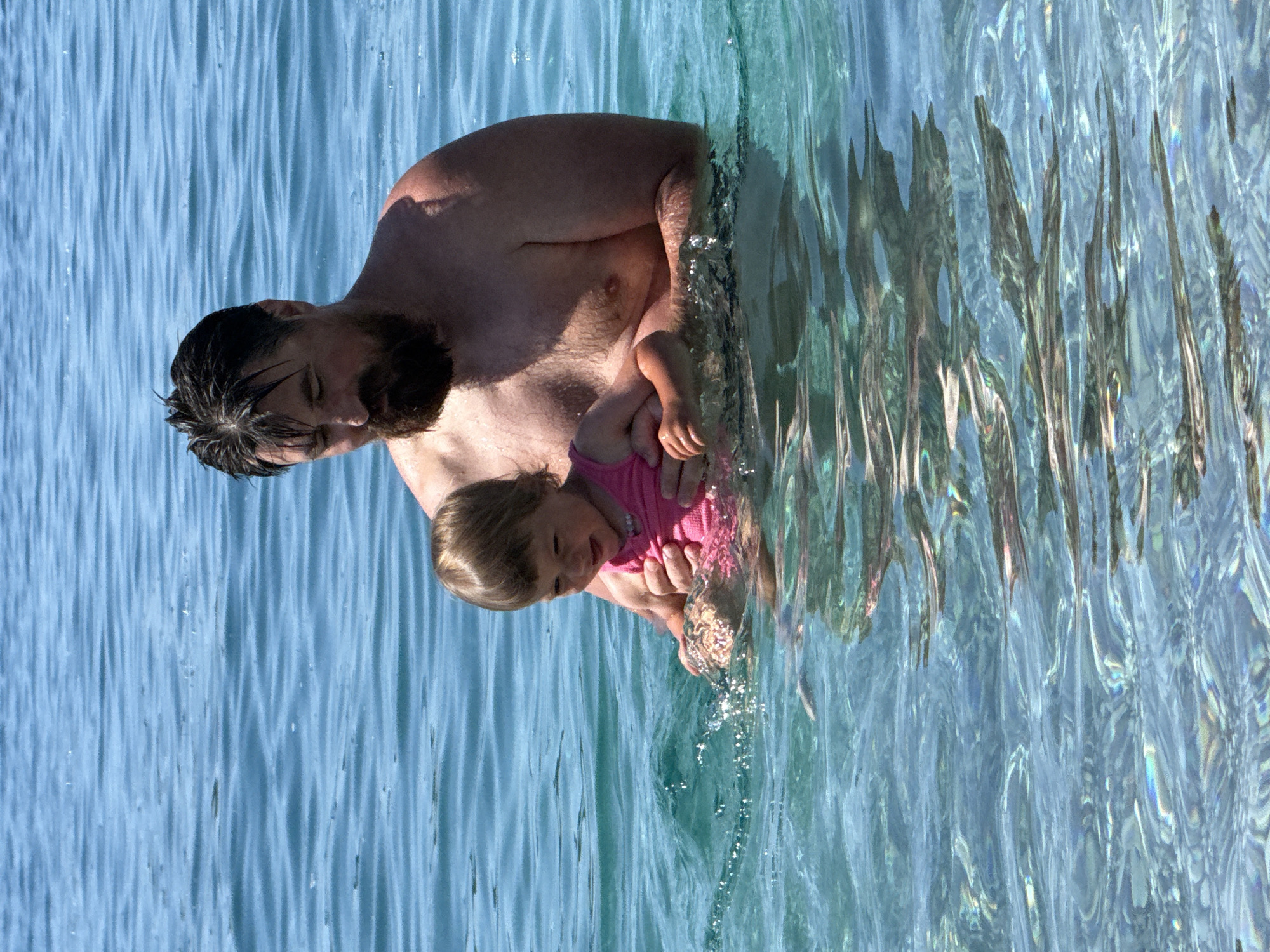 The image displays an adult male and a young female child standing chest-deep in clear, turquoise water under bright sunlight in Boreti, Montenegro. The adult male has dark, wet hair and a prominent dark beard; his eyes are directed downwards or closed. He is bare-chested and holds the child with both hands, supporting her in front of him. The child, a toddler with light brown hair, wears a pink top or swimsuit and exhibits a smiling facial expression, looking towards the water disturbed by the adult's arm. The water's surface is lightly rippled with small air bubbles visible around the child and the adult's hands. The water is highly transparent, revealing light refraction patterns on the submerged areas. The background consists entirely of open water, appearing a darker blue further from the subjects.