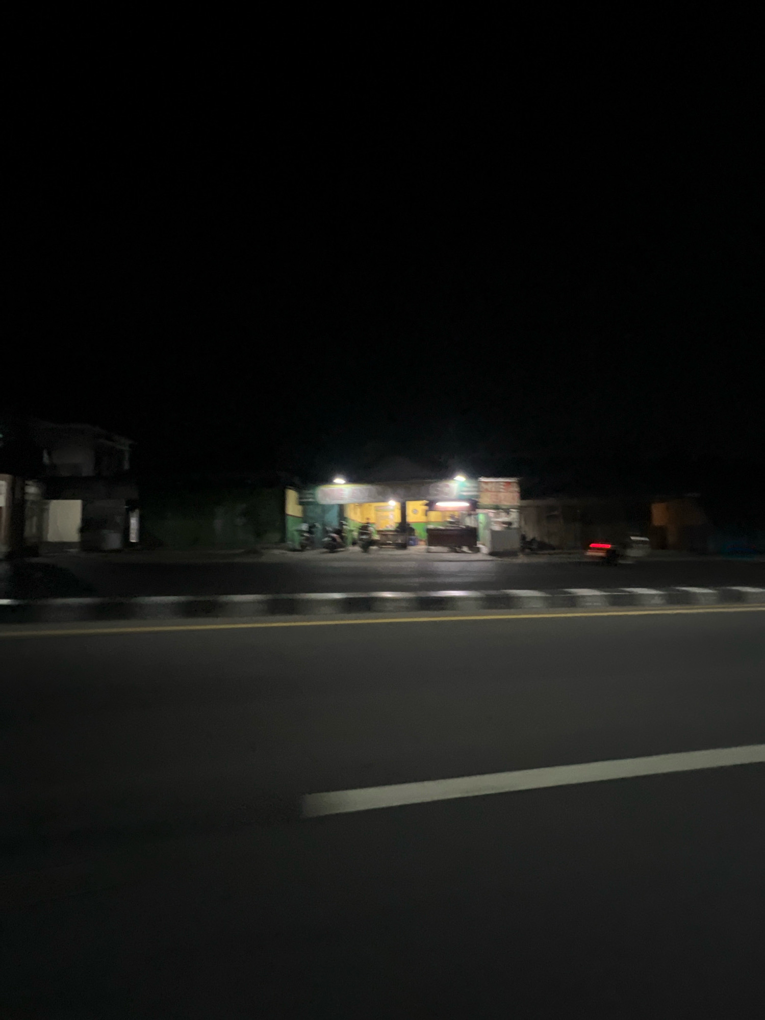 The image depicts a nighttime scene on a road in Sucen, Indonesia, likely in the vicinity of roadside businesses. The foreground is dominated by a dark asphalt road with a white dashed line and a yellow solid line marking the lanes. Beyond the road, a row of dimly lit structures suggests small shops or food stalls. Several motorcycles are parked in front of these establishments. One structure, appearing to be a simple building, is visible on the left. A red light, possibly from a vehicle, is visible in the middle ground on the right. The sky is completely dark, indicating it is late at night. The overall impression is one of a quiet, sparsely lit commercial area along a thoroughfare.