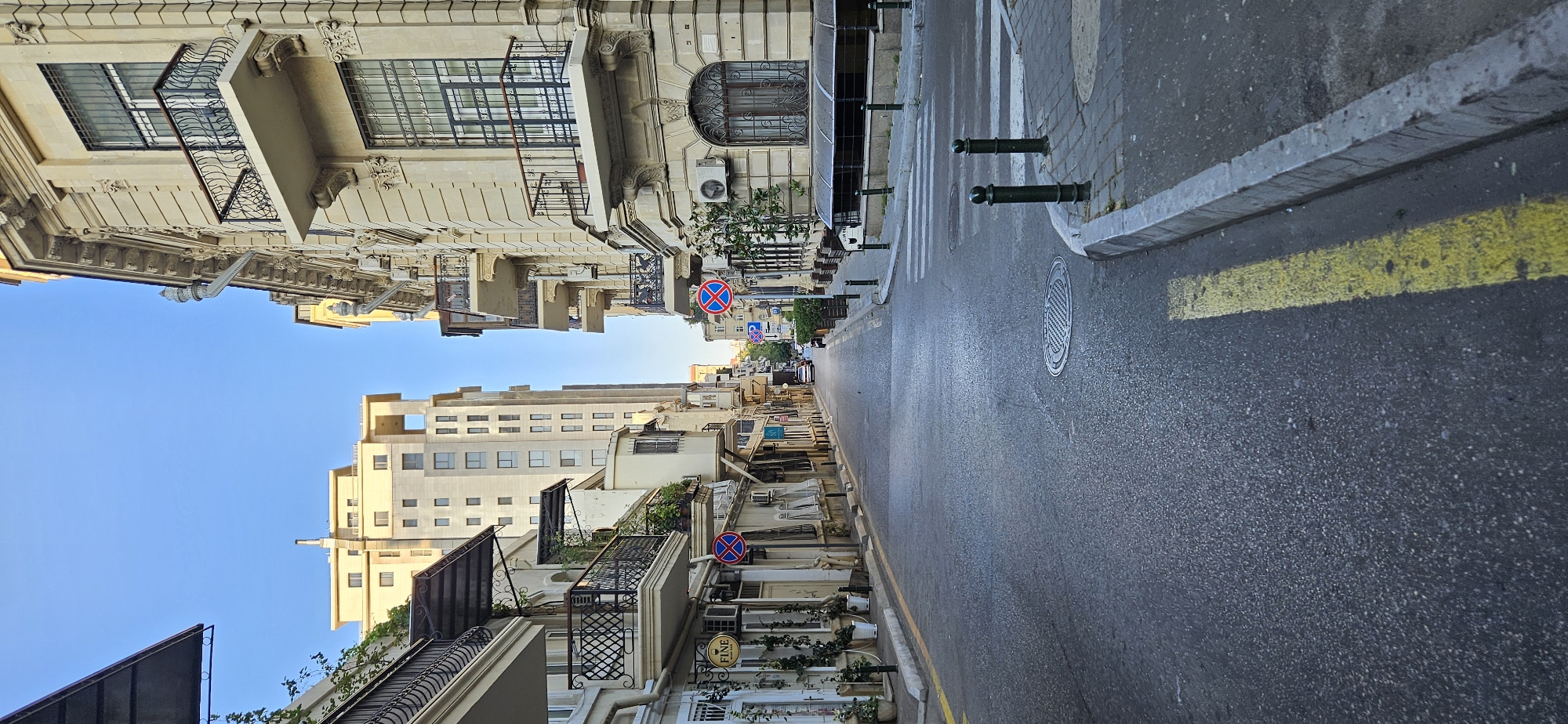 This is a vertical, eye-level, medium shot of a street in the city of Baku, Azerbaijan, taken on a clear day. The perspective is looking down a relatively narrow asphalt street flanked by buildings. On the left, a building with balconies and awnings is visible. A circular sign with a red border and a red diagonal line through a blue circle is mounted on the building. On the right, a grander, ornate building with multiple balconies, intricate stonework, and arched windows is prominent. This building also has a similar circular sign with a red border, red diagonal line, and a blue circle. Further down the street, other buildings of varying architectural styles are visible, including a tall, modern high-rise in a pale color. The street has white pedestrian crossing markings and a yellow line marking the edge of the road. There are bollards and manhole covers on the street. The sky is clear and blue. There are no people or vehicles clearly visible in the immediate foreground.