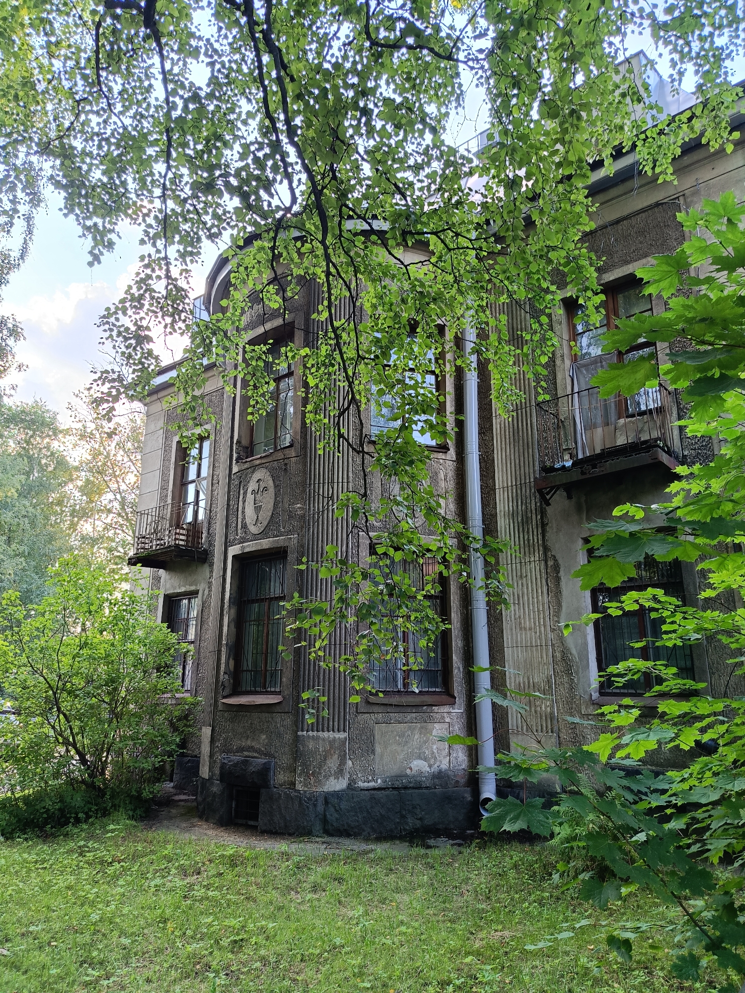 The image shows the exterior of a weathered, old, multi-story building with an ornate facade, partially obscured by lush green trees and foliage. The building appears to be made of stone and has several windows, some with balconies and metal grates. A vertical pipe runs down the side of the building. The ground in front of the building is covered with green grass. The overall atmosphere suggests a somewhat overgrown and possibly abandoned property in a city setting.
