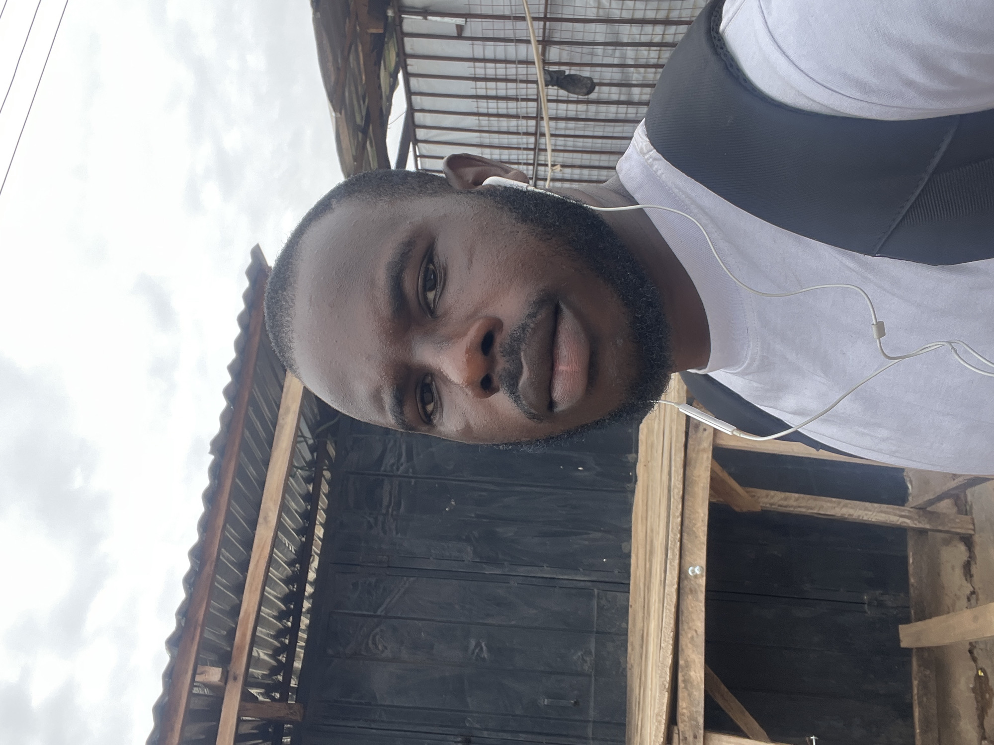 A man with dark skin, a full beard, and a white t-shirt with a black strap over his shoulder is taking a selfie. He is wearing wired earphones. The background shows a structure of corrugated metal sheets, black painted wooden panels, and wooden beams. This is in Jos, Nigeria.