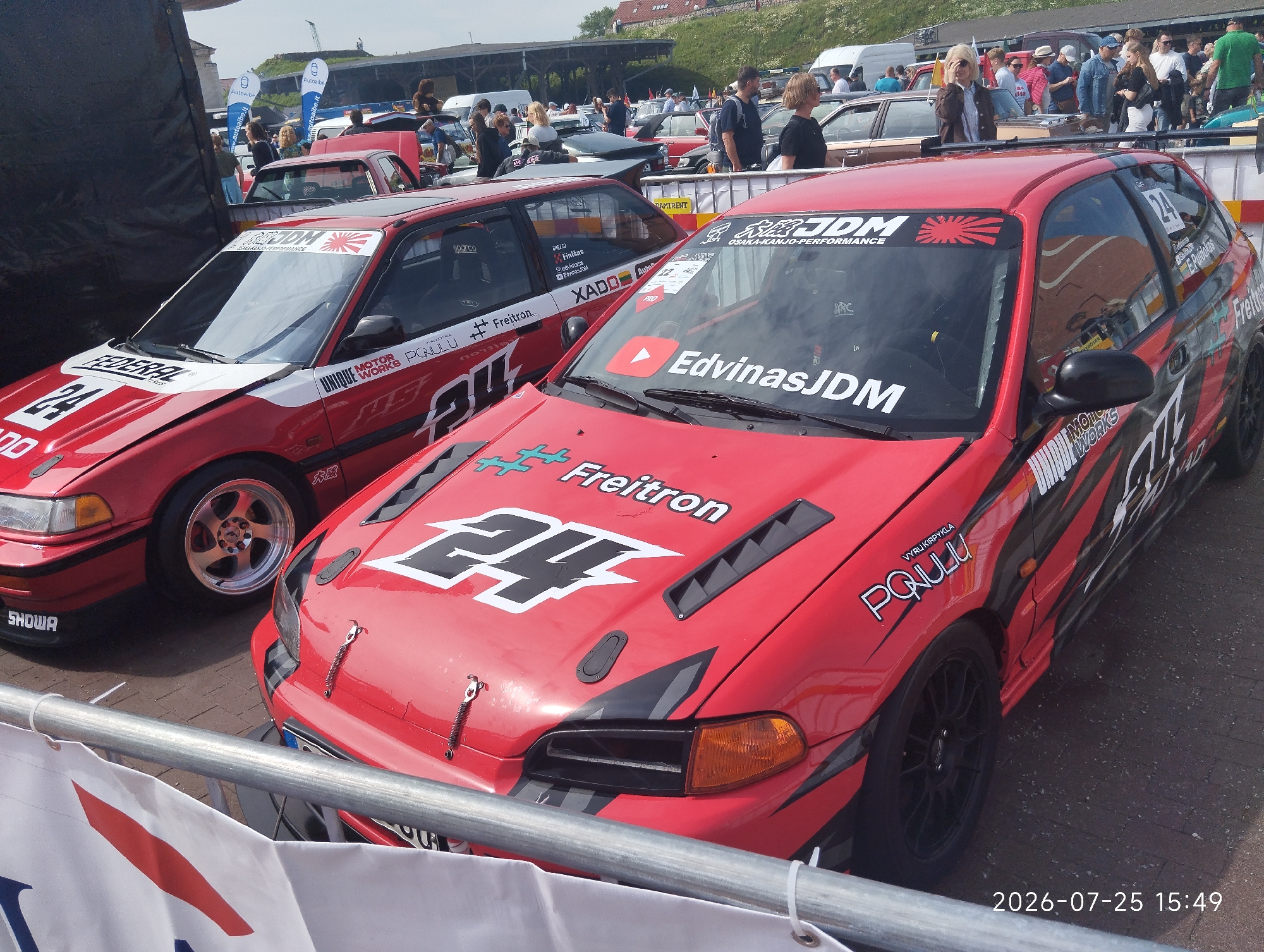 The image depicts a street event, likely a car show or race, with several modified red cars parked on an asphalt surface. In the foreground, two red hatchback cars are prominently displayed. The car on the left has "FEDERAL" and "SHOWA" decals, with a large "24" and "MONTE WORKS" text on its hood and side. The car on the right features a "JDM" decal on its windshield, a YouTube play button logo, and "EdvinasJDM" text. Its hood has a "24" graphic and "Freitron" branding. Several people, dressed casually, are visible in the background, observing the cars and the event. The environment appears to be outdoors, possibly a parking lot or a closed-off street, with some fencing in the foreground. In the far background, there are buildings and possibly a hill. The overall atmosphere suggests a sunny day and a public gathering focused on automotive culture. The timestamp "2026-07-25 15:49" is superimposed on the bottom right corner. The text "MIESTO automobilių sporto festivalis KLAIPĖDA 2024" visible on a banner in the background places the event in Klaipėda, Lithuania, in 2024.