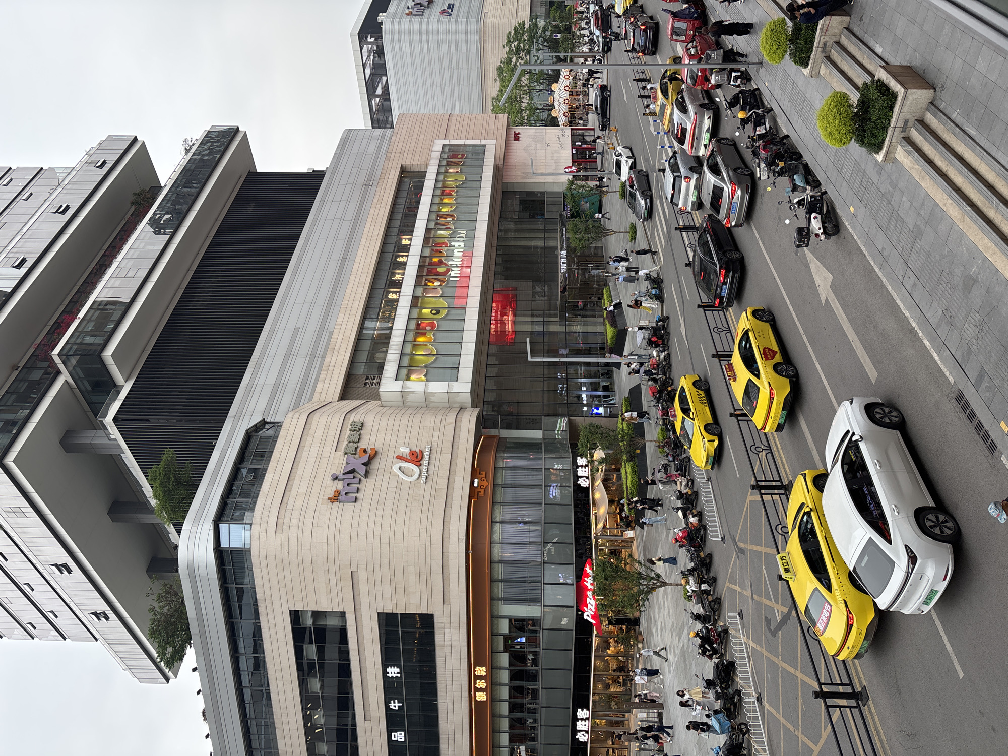 This eye-level, outdoor photograph depicts a bustling street scene in the Jiulongpo District of China. The street is filled with pedestrians, parked motorcycles, and a line of taxis with a color scheme of yellow and white. Buildings line the street, the most prominent being a multi-story shopping center complex with visible retail stores such as Pizza Hut, Ole Supermarket, and brands written in Chinese characters. A modern high-rise building with a stepped facade stands beside the shopping center. The sky is overcast.