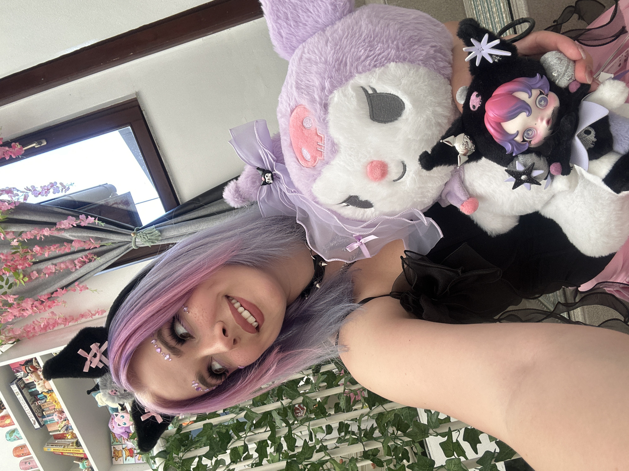 A young woman with pink and lavender hair wears black cat ears and facial gems, holding a large purple plush toy and a smaller black-and-white plush toy with a character head. She smiles at the camera. The setting includes a bookshelf, window with curtains decorated with pink flowers, and artificial green foliage. The location is unclear, as there are no specific indicators of the City of Zagreb, Croatia.