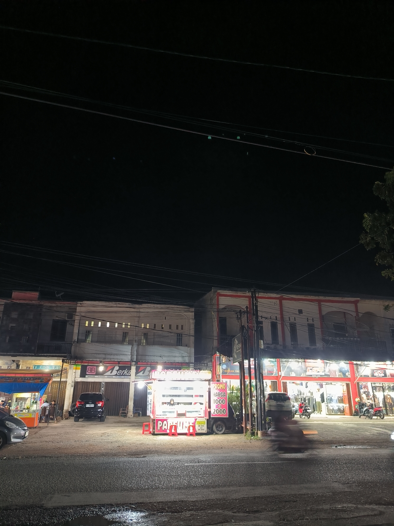 The image captures a bustling nighttime street scene in Banda Aceh, Indonesia, illuminated by artificial lights against a dark sky. Several multi-story commercial buildings line the road, housing various businesses. In the foreground, a wet asphalt road shows reflections from the lights, suggesting either recent rain or dampness.
On the left side of the street, a blue vendor stall is visible, advertising "Zakey Juice" and "MAKRIDE KUAG MERAM." A grey sedan is partially seen in front of it, and a few indistinct figures are near the stall. Next to it, a darker building features a sign with "CV. JEMBAR LOGAM" and another for "Berka B DESI," with a black Toyota Innova (license plate P 1855 JI) parked directly in front.
In the center, a brightly lit white food or merchandise cart with a red base prominently displays a "PROMO HARGA 1000 S/D 2000" sign, along with "PARTUME" (likely Parfum) and another "PROMO RP. 1000" sign. To its right, a larger building with red and white trim showcases a brightly lit storefront, likely a clothing store, with "OCEAN PAC" partially visible on a sign above. Several motorbikes are parked in front of this shop.
A blurred figure on a motorbike is captured in motion on the road, indicating ongoing traffic. Numerous overhead electrical wires crisscross the dark sky, a common feature in the urban landscape. The scene conveys the active, commercial atmosphere of a street in Southeast Asia after dark.