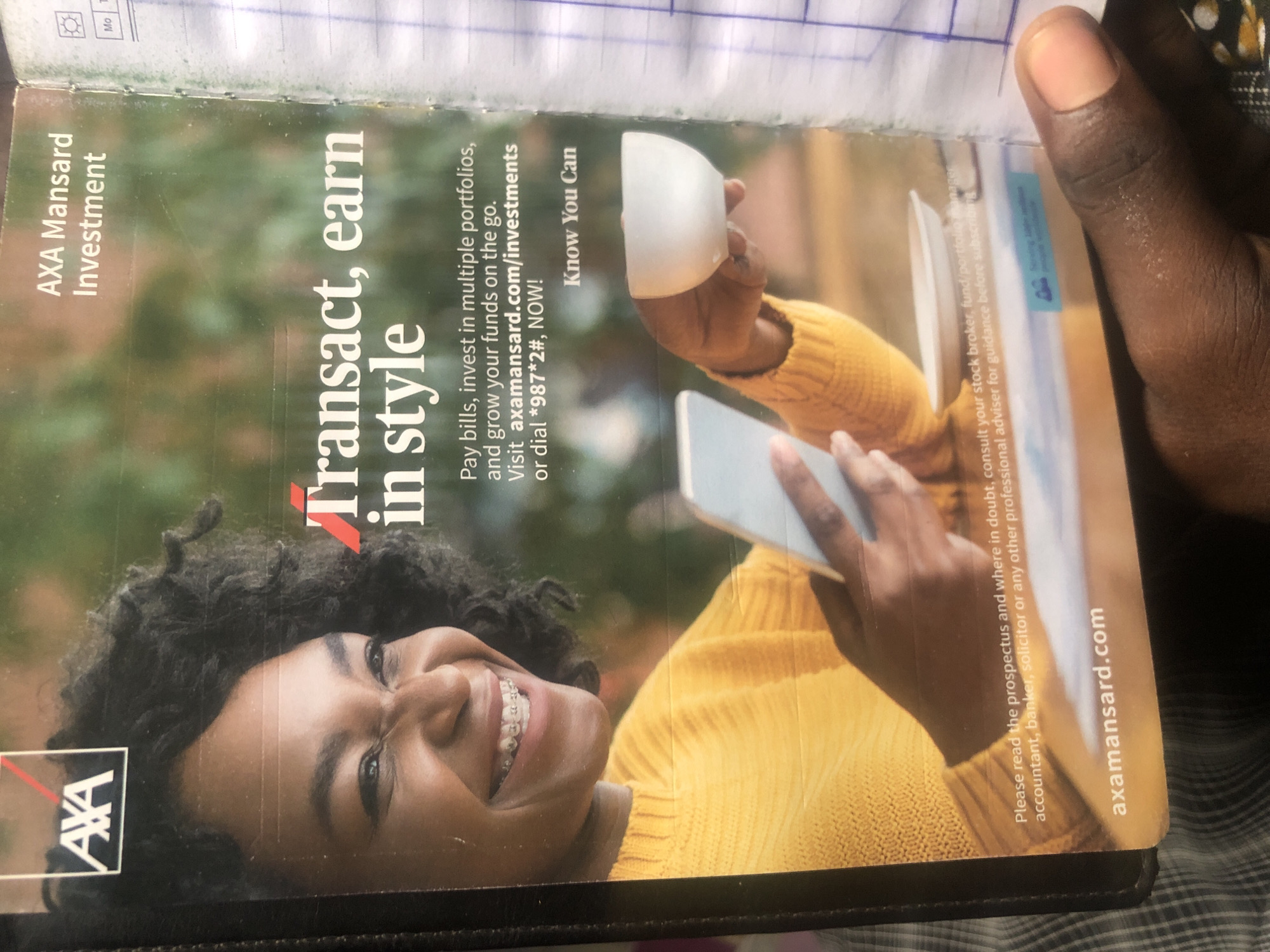 This is a close-up, high-angle, outdoor photograph of a magazine advertisement for AXA Mansard Investment. The advertisement features a smiling Black woman with curly hair and braces, wearing a yellow knitted sweater. She is holding a smartphone in her left hand and a small white bowl in her right hand. The background is a blurred green foliage. Text overlays the image, including the AXA logo, "AXA Mansard Investment," and the tagline "Transact, earn in style." The bottom of the advertisement includes a disclaimer about reading the prospectus and consulting professional advisors, along with the website "axamansard.com." The photograph is taken from a perspective that includes the edge of a black binder and a person's thumb in the lower right corner. This image is not specific to Ado, Nigeria, and provides no information about the city.