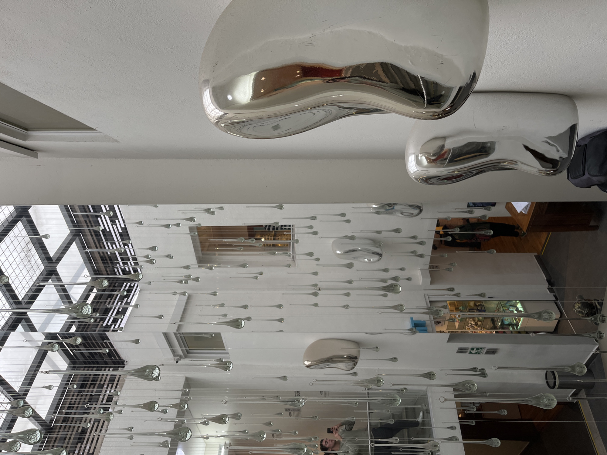 The image shows an interior scene in Mexico City, Mexico, featuring a large, modern chandelier-like installation with numerous small, teardrop-shaped glass orbs suspended by thin wires from the ceiling. The wires create a dense, three-dimensional effect, filling the space between the ceiling and the floor. Two irregular, silver-colored light fixtures are affixed to the ceiling, reflecting the interior environment. The architecture includes a gridded skylight, and reflections of the interior are visible on the glass orbs. A person appears to be taking a photograph of the chandelier with a smartphone. Elements of the background suggest the setting is a gallery or public space, evidenced by displays and furnishings.