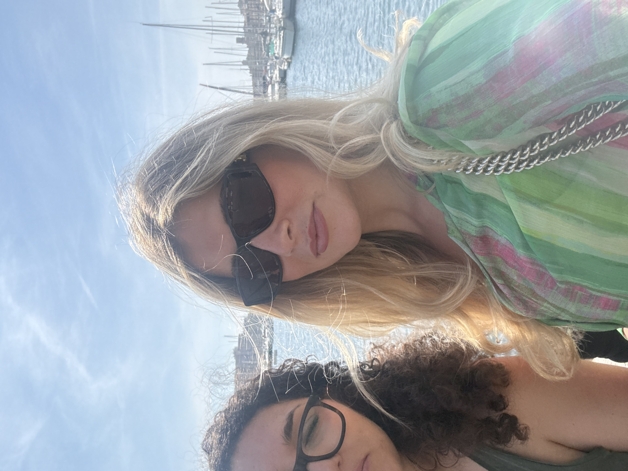 An informal portrait captures two women posing outdoors, likely at a marina in Marseille, France. The image is oriented with the subjects' heads tilted to the left, as if taken from a vertical phone held horizontally.
In the foreground, a woman with long, flowing blonde hair, styled with subtle waves, wears dark sunglasses and a light green, possibly sheer, patterned top with hints of pink and other colors. A silver chain accessory is visible on her shoulder. She has a relaxed expression with slightly parted, pinkish lips, and her face is partially illuminated by direct sunlight.
Behind her, partially visible on the bottom left, is another woman with dark, curly hair and dark-rimmed glasses. Only the upper part of her face, including her eyes, and a shoulder in a dark top are visible, suggesting she is slightly behind and to the side of the first woman.
The background reveals a bustling marina under a bright, clear blue sky with minimal wispy clouds. Numerous sailboat masts and the tops of various boats and yachts are visible, hinting at the coastal setting. The calm, blue water of the harbor is also present. The scene is well-lit, indicating a sunny day, likely morning or afternoon, fostering a bright and pleasant atmosphere. No visible text is present in the image.