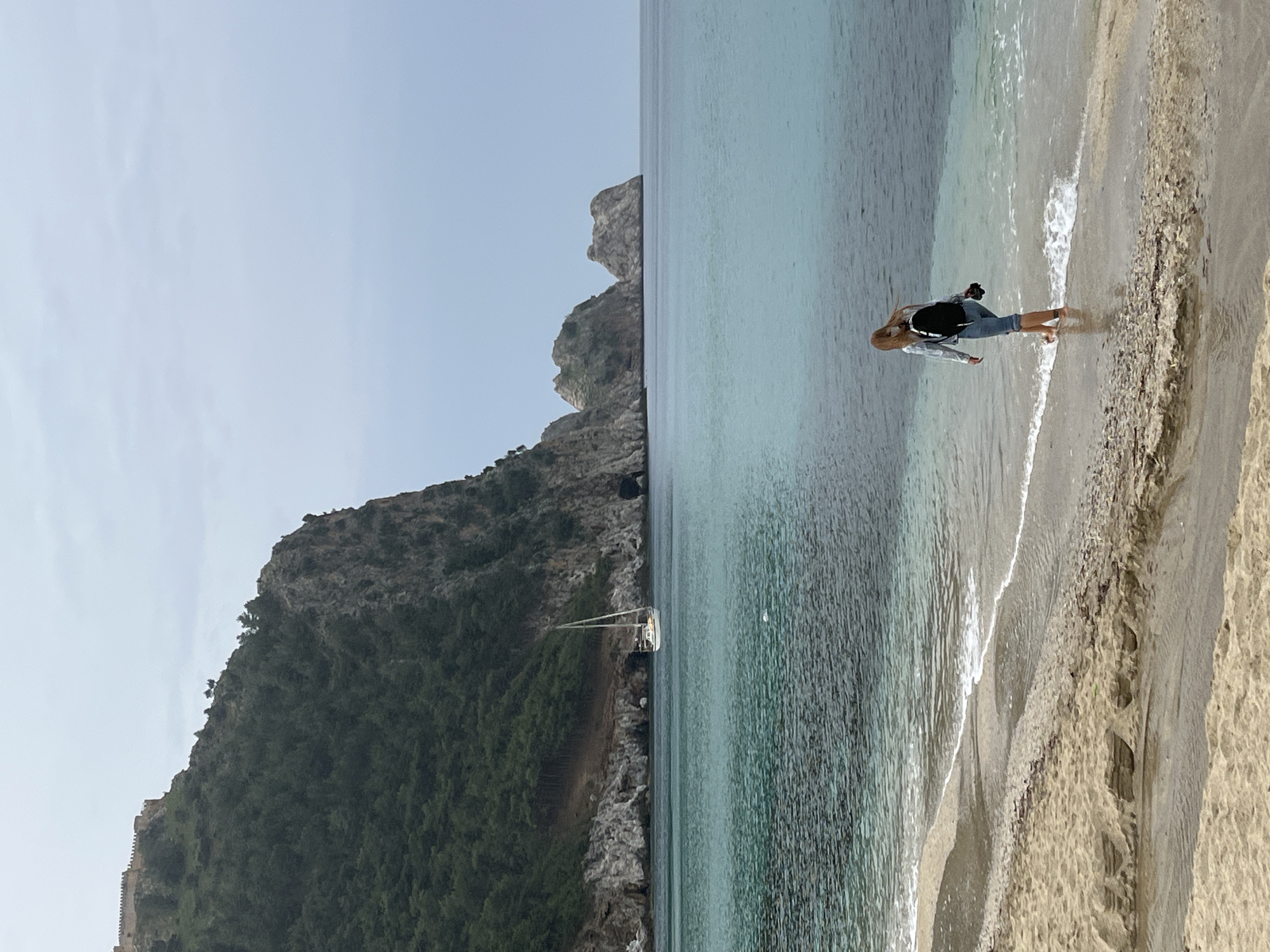 The image captures a tranquil coastal scene in Alanya, Turkey, featuring a person on a beach, the sea, and a dramatic mountainous backdrop.
In the foreground to the right, a woman with blonde hair, dressed in a light-colored long-sleeved top (possibly a light jacket or denim shirt), a dark top underneath, and light-colored pants, stands barefoot at the edge of the water. She appears to have a camera or binoculars hanging around her neck. She is positioned with her back partially to the viewer, looking down towards the gentle waves lapping at her feet on the sandy and pebbly shore. Her posture suggests she is either observing something in the water or perhaps taking a photo.
The sea, a clear turquoise-blue, extends across the middle and right side of the frame, showing gentle ripples and small white waves breaking near the shore. Further out in the water, positioned closer to the mountain, a small white sailboat or yacht is anchored.
Dominating the left side of the scene is a massive, dark green, tree-covered rocky mountain or cliff face. Towards the base of this mountain on the far left, remnants of an ancient wall or fortification can be seen. Another lighter, more barren rocky outcrop is visible jutting out from the mountain into the sea, further in the distance.
The time of day appears to be daytime, with clear, bright conditions and a light blue sky, indicating pleasant weather. The overall impression is one of natural beauty and peaceful enjoyment of the seaside environment. There is no visible text in the image.