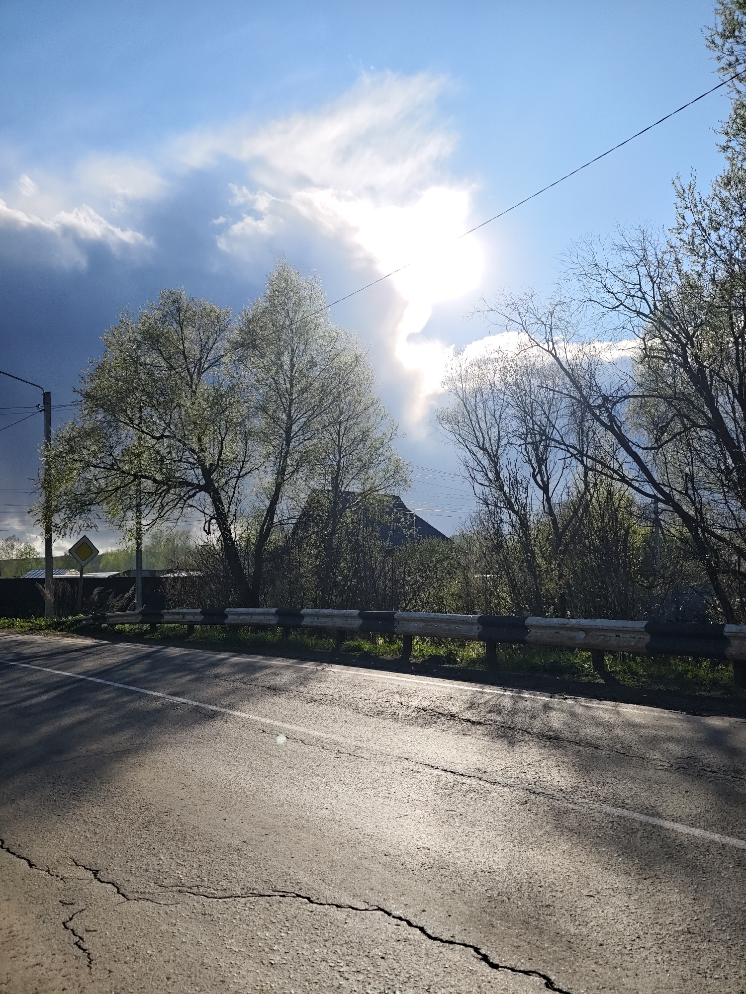 The image depicts a road in Malyi Bukor, Russia. The asphalt road surface has cracks and a white line marking. A guardrail is positioned along the roadside. Beyond the guardrail, there are trees and vegetation. A road sign is visible. In the background, a building roof can be seen. The sky is partly cloudy with a bright sun. Overhead wires are present.