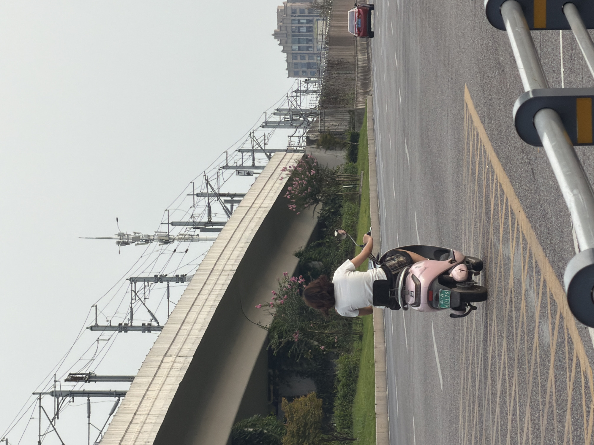 The image shows a woman riding a pink electric scooter on a road in Jiangjin District, Chongqing, China. She is wearing a white t-shirt and dark shorts, with her back to the camera. The scooter has a license plate reading "6D179." In the background, an elevated highway with overhead power lines is visible, along with some buildings and vegetation. A red car is also seen on the road ahead. The sky is overcast.