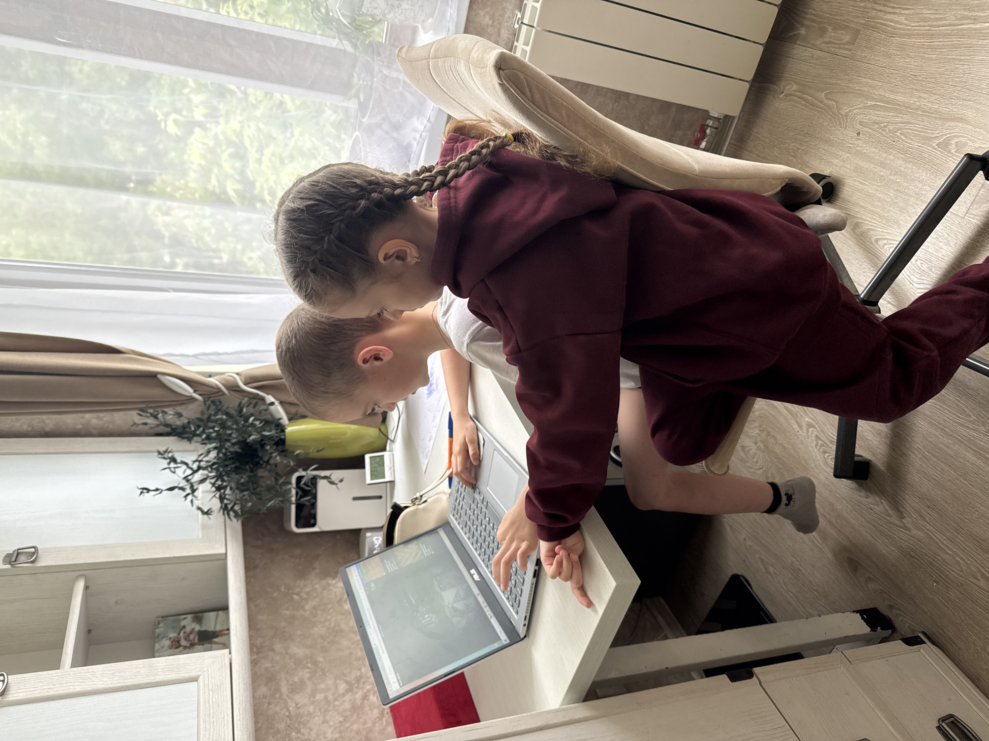 Two children are looking at a laptop. A girl with braided hair is wearing a burgundy hoodie and is leaning over a boy who is sitting at a desk. The desk is located near a window. The room has wooden floors and light-colored furniture. The location is Razdolie, Russia.