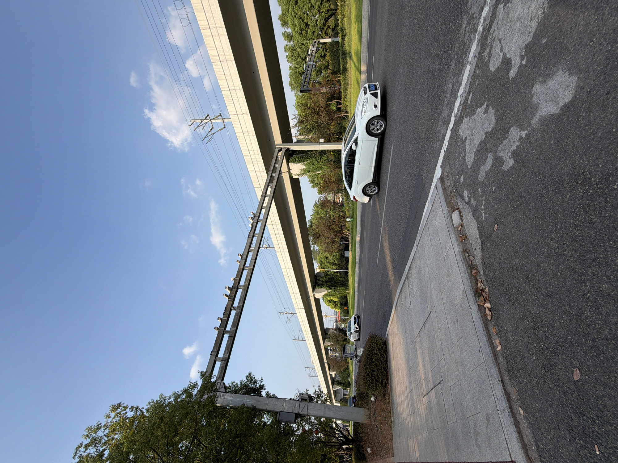 The image shows a wide, paved road with a sidewalk on the left side, bordered by a tree line and a large concrete structure supporting overhead electrical wires, possibly for a train or light rail system. A white sedan is driving on the right side of the road, heading away from the camera. The sky is blue with scattered white clouds. The scene appears to be in an urban or suburban area. This location is identified as Jiangjin, China.