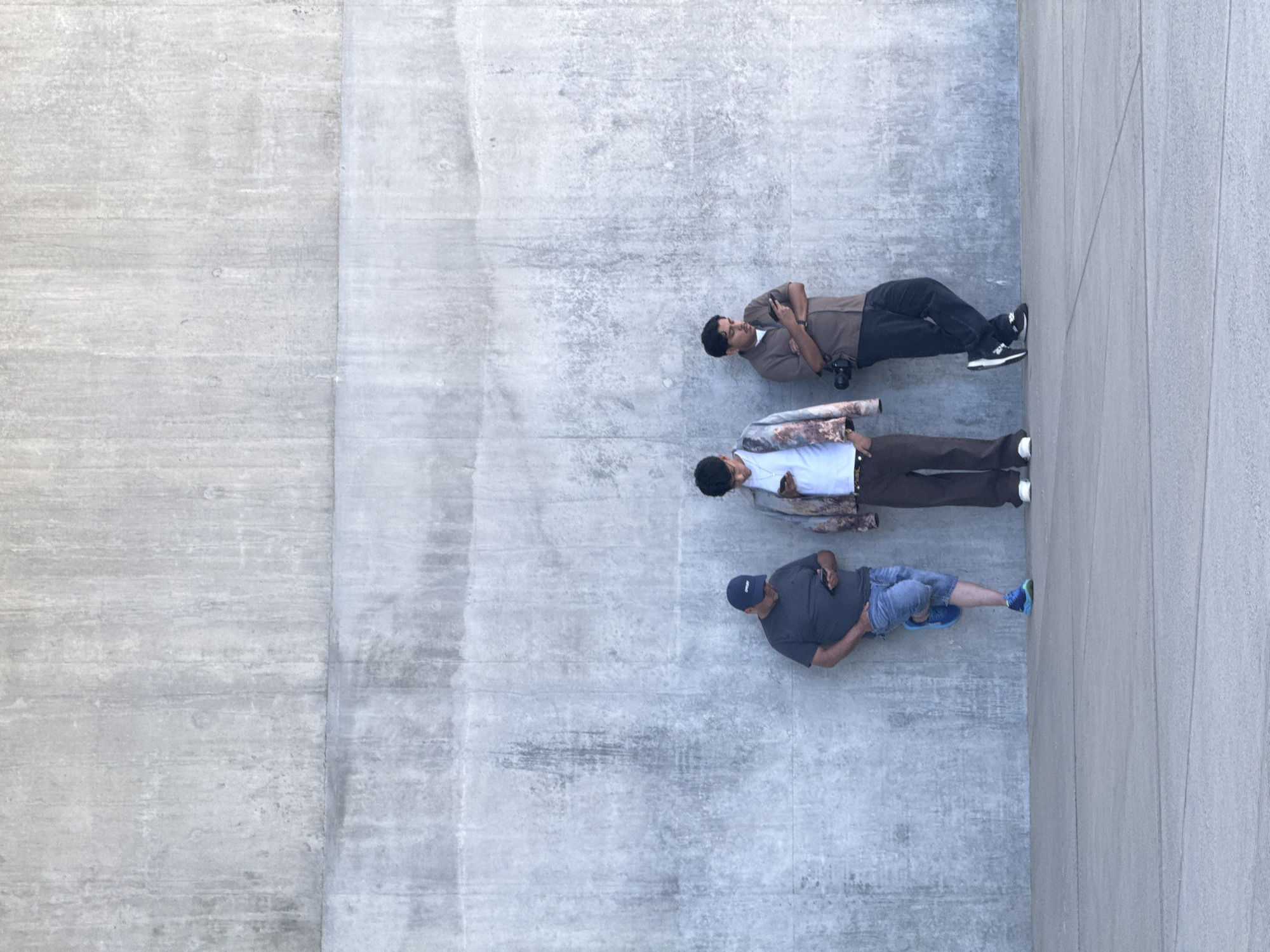Three men stand against a large, textured concrete wall. The man on the left wears a dark grey t-shirt, blue denim shorts, and blue sneakers, with one leg crossed over the other. He is looking down at his phone. The middle man is dressed in a light blue t-shirt, brown patterned jacket, and brown pants, also looking at his phone. The man on the right wears a brown short-sleeved shirt, dark pants, and black and white sneakers. He has a camera hanging from his shoulder and is also looking at his phone. The ground is a flat, light grey concrete surface. The scene is an outdoor urban environment. This image was captured in Los Angeles, United States of America.