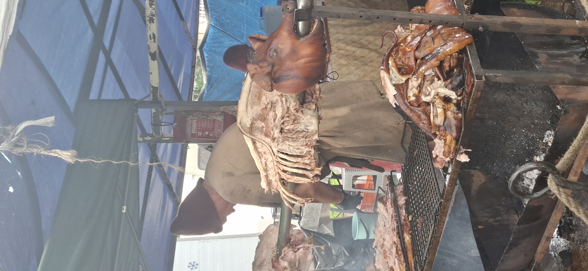 The image shows a pig being roasted on a spit, outdoors, under a blue and green tarp. A person wearing a brown conical hat and a tan apron is standing near the spit, attending to the pig. The pig is whole, with its head and body visible. The meat is browning and looks cooked. There are also other pieces of meat on a grill below the spit. A sign on the tarp reads "SAMMUTUSPEN BRANDFIL". The scene appears to be at an outdoor market or festival. The overall impression is of a food vendor preparing and selling roasted pork.