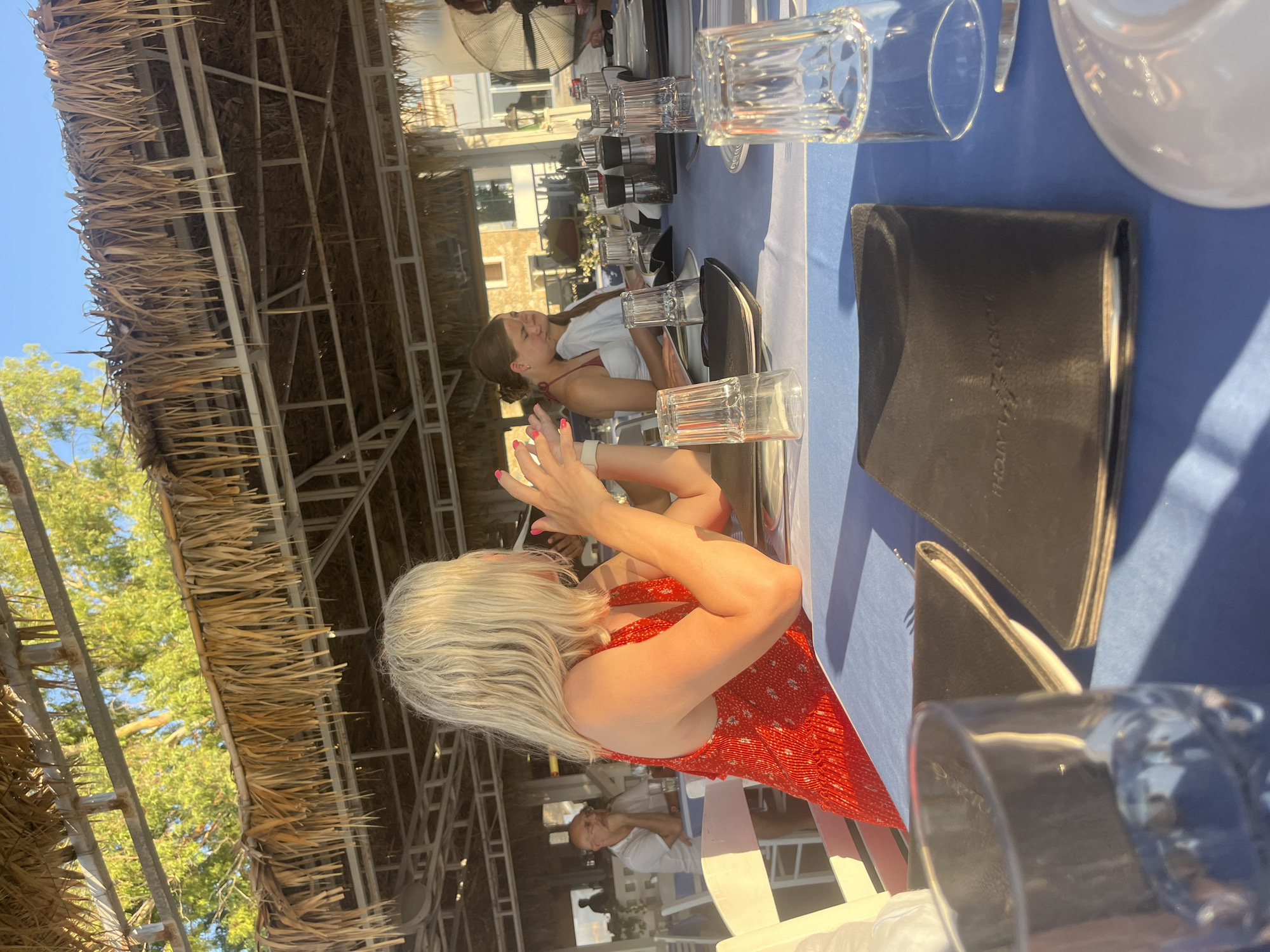 The image depicts a group of people seated at a table on a patio, likely a restaurant, with a thatched roof overhead. The setting appears to be outdoors, illuminated by natural sunlight, casting distinct shadows. In the foreground, a woman with blonde hair, wearing a red dress, is seated at the table, her hands raised as if speaking or gesturing. She is looking to her right. Across from her, a young woman with brown hair, wearing a white top, is also seated at the table. Further back, a man with glasses and a white shirt is visible, seated at another table. The table is set with blue tablecloths, napkins, menus, silverware, and glassware. In the background, trees and buildings are visible, suggesting an urban or semi-urban environment. The overall atmosphere is casual and relaxed, indicative of a meal or gathering. Based on the presence of palm trees, the blue tablecloths, and the style of the buildings in the background, this scene is likely located in Polis Chrysochous, Cyprus.